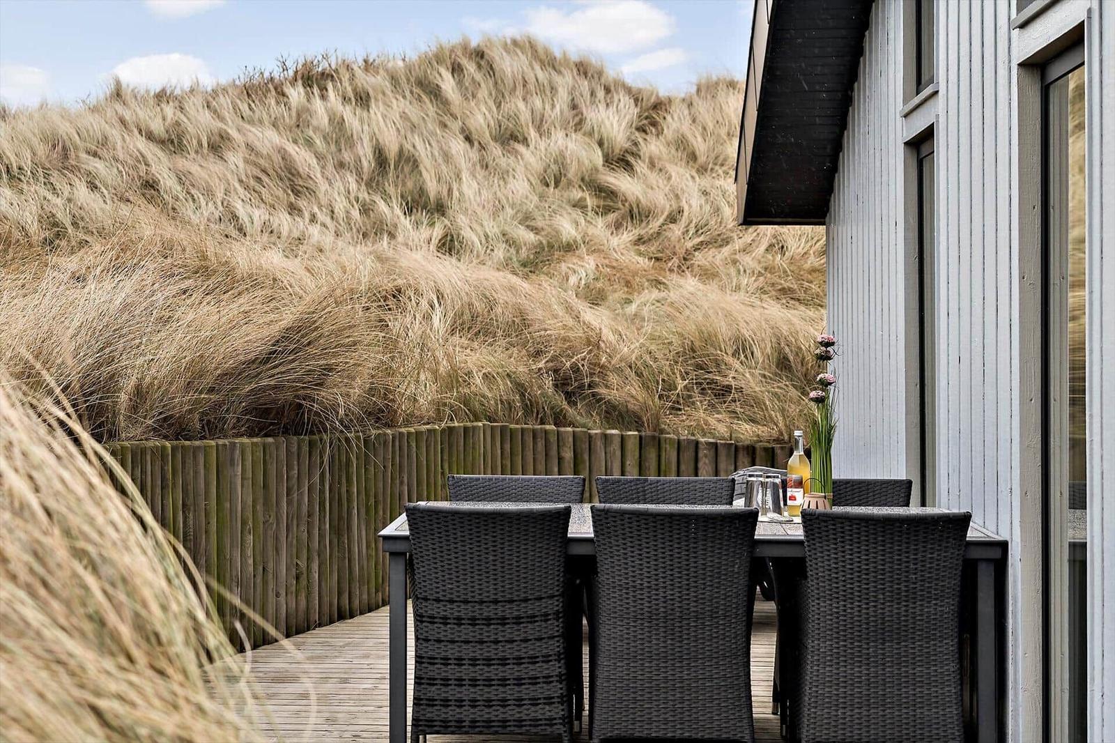 Terrasse mit Tisch und Stühlen neben einem Haus mit Dünen im Hintergrund.