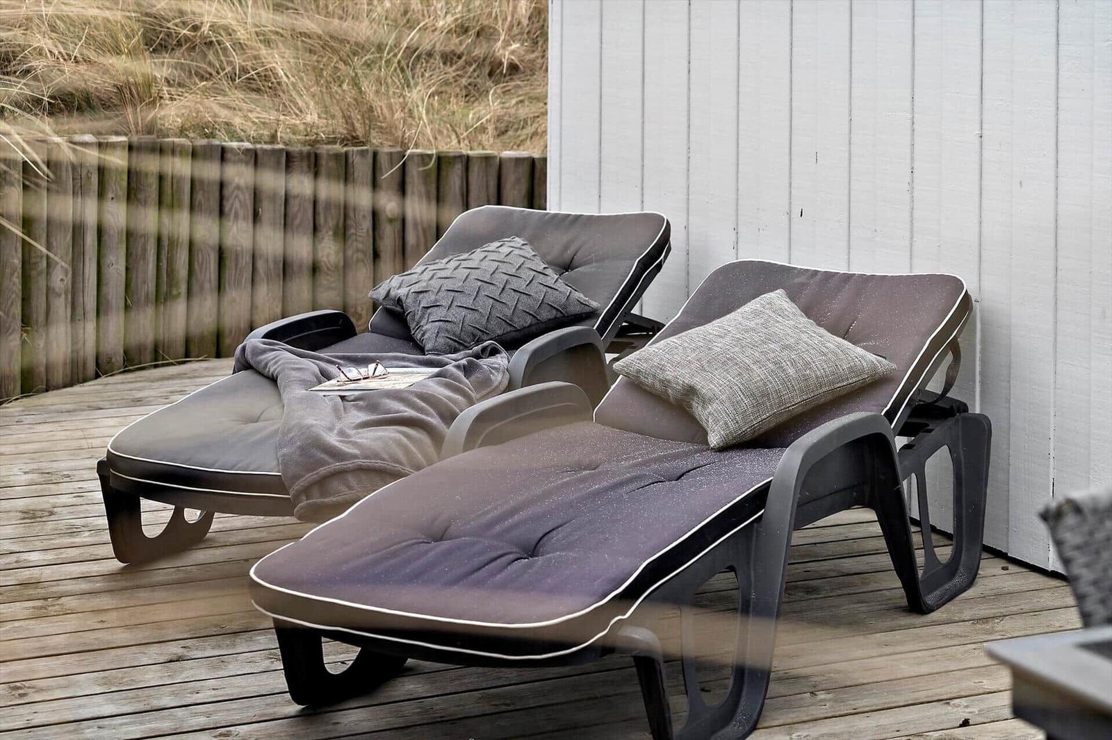 Zwei Liegestühle mit Kissen und Decke auf einer Holterrasse neben einer weißen Wand.