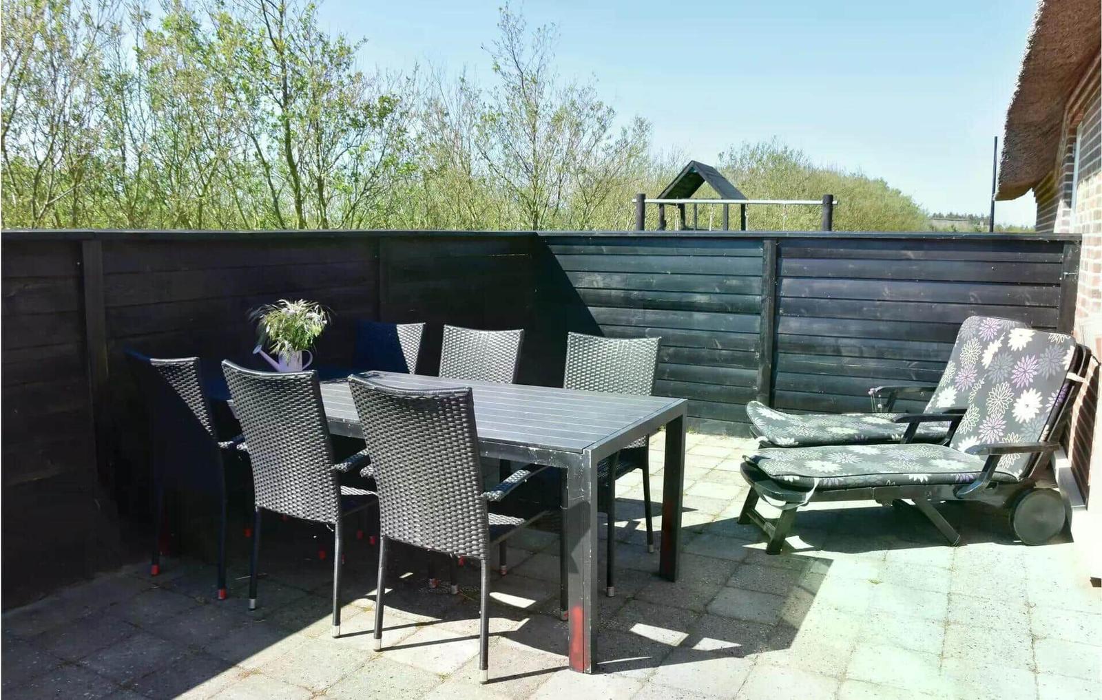 Terrasse mit Tisch, Stühlen und Liegestühlen. Hintergrund: Holzzaun und Bäume.