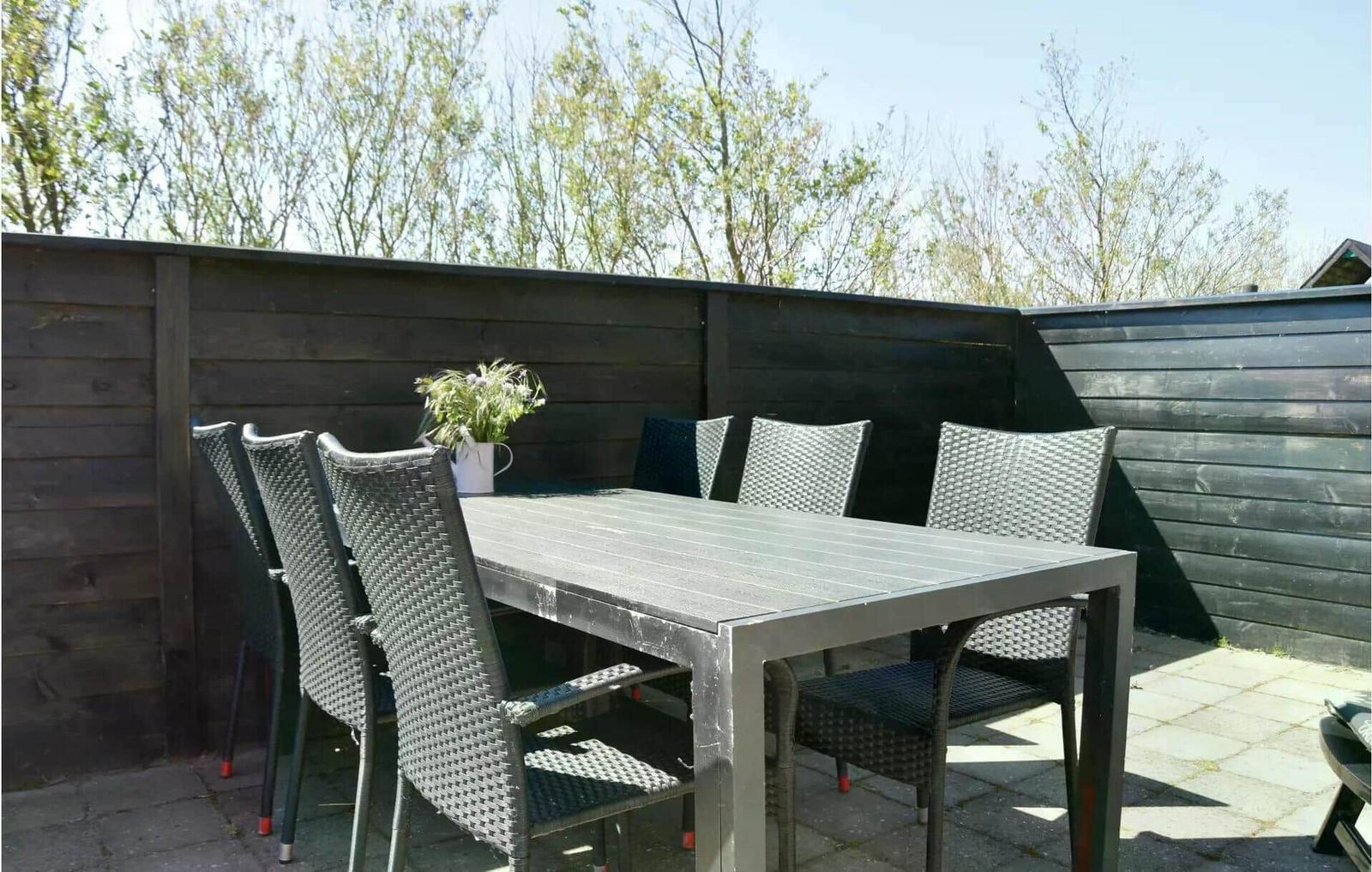 Terrasse mit Tisch und sechs Stühlen, umgeben von dunklem Holzzaun und Bäumen.
