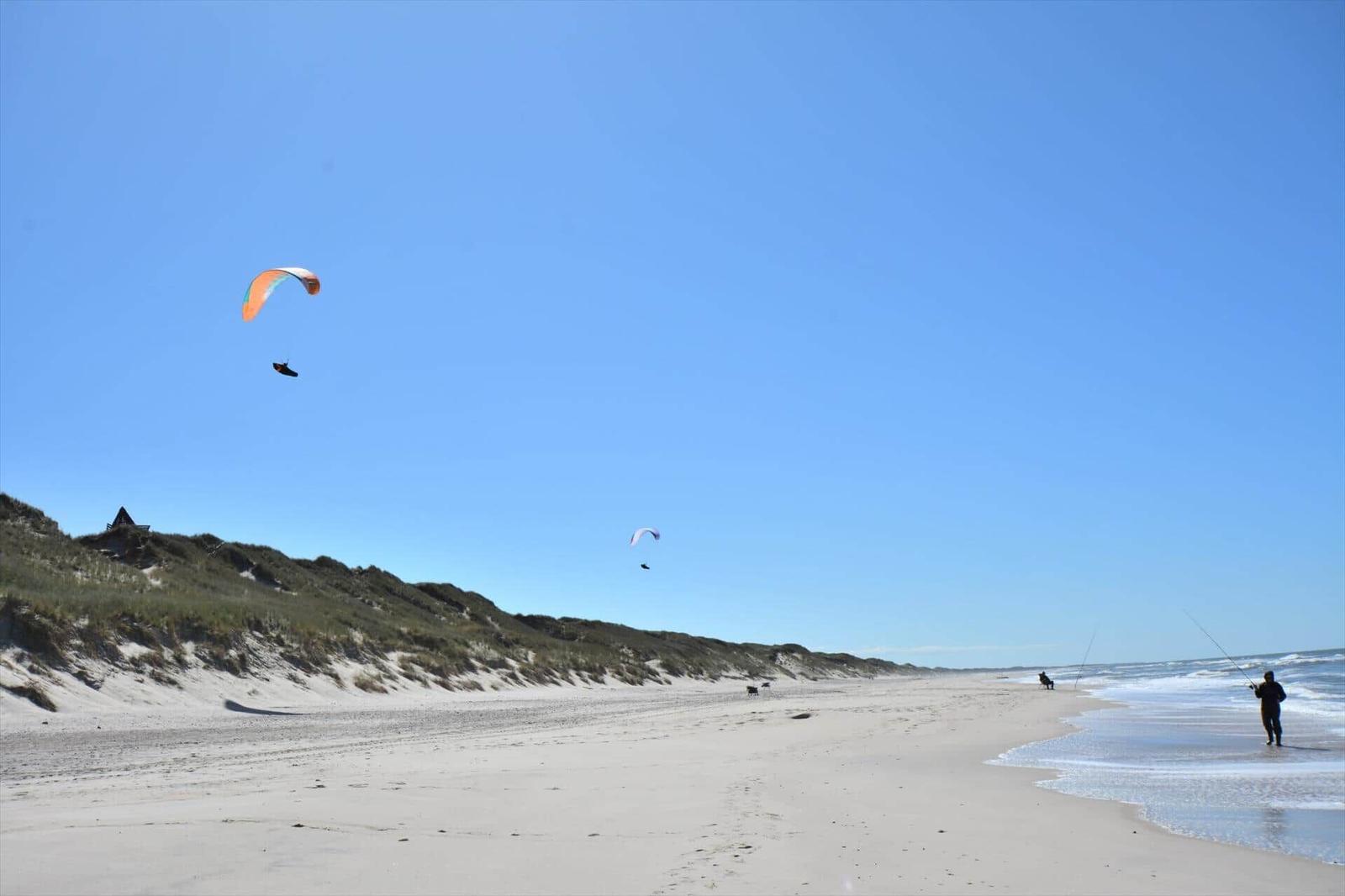Strand mit Sanddünen und Gleitschirmfliegern am Horizont.