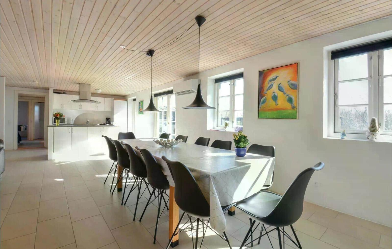 Essbereich mit langem Tisch und Stühlen, Küche im Hintergrund, Holzdecke und Fenster.