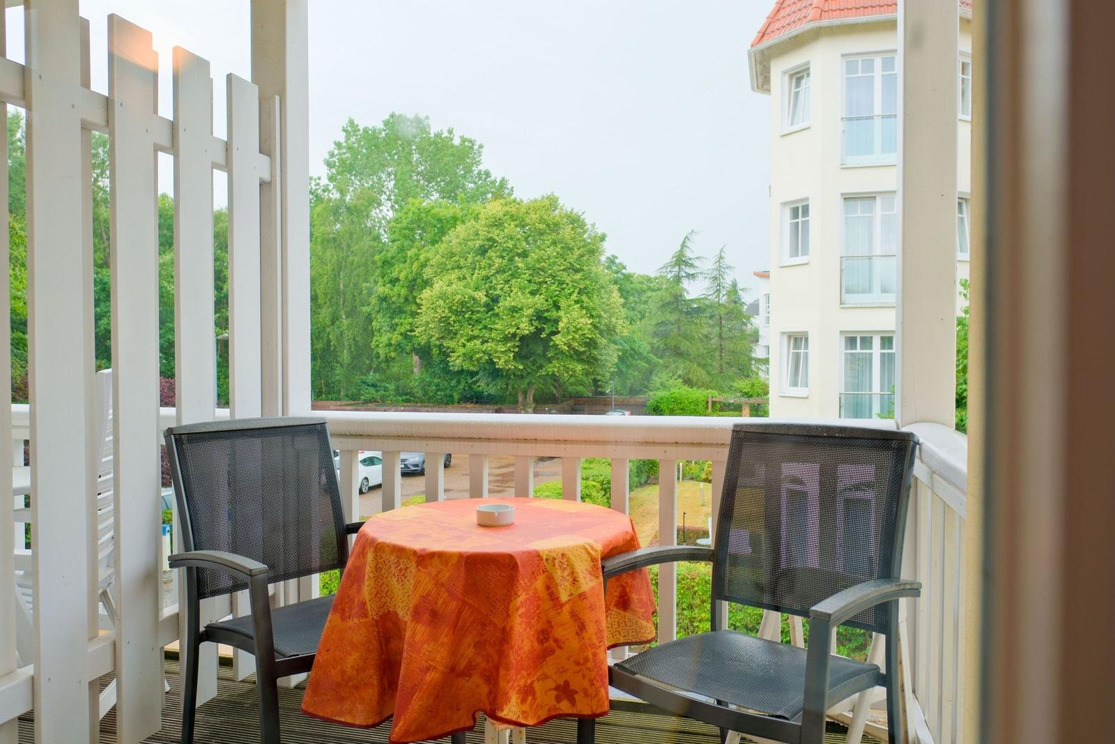 Balcony with table and two chairs, covered with orange tablecloth, overlooking green trees and building.