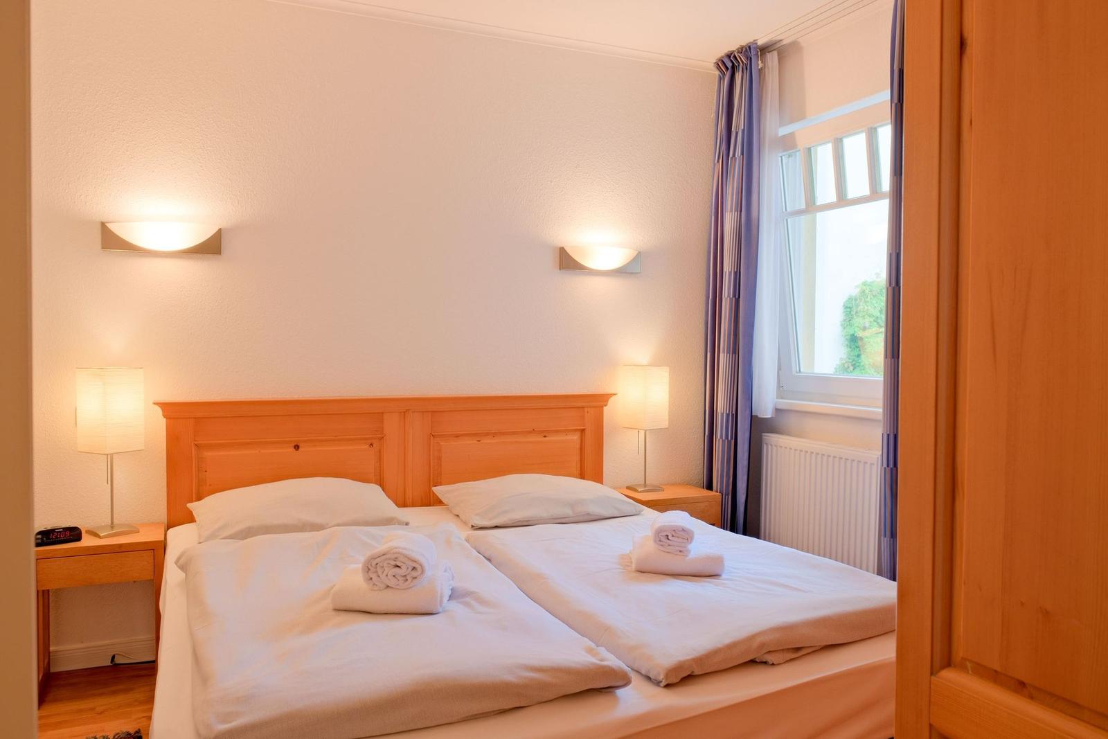Doppelbett mit weißen Laken, zwei Nachttische und Fenster mit Vorhängen.