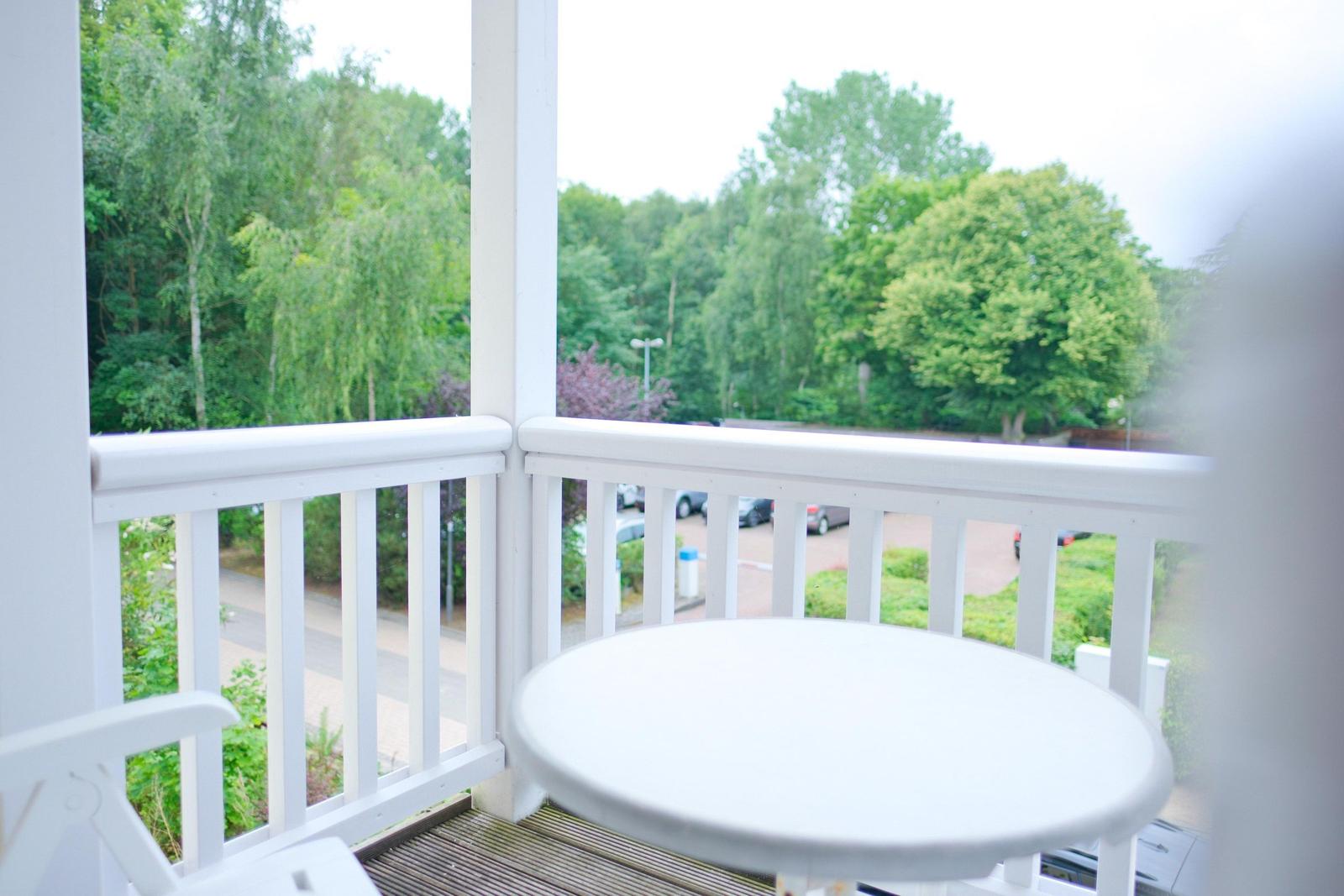 Terrasse mit weißem Tisch und Stühlen. Blick auf grüne Bäume und Parkplatz.