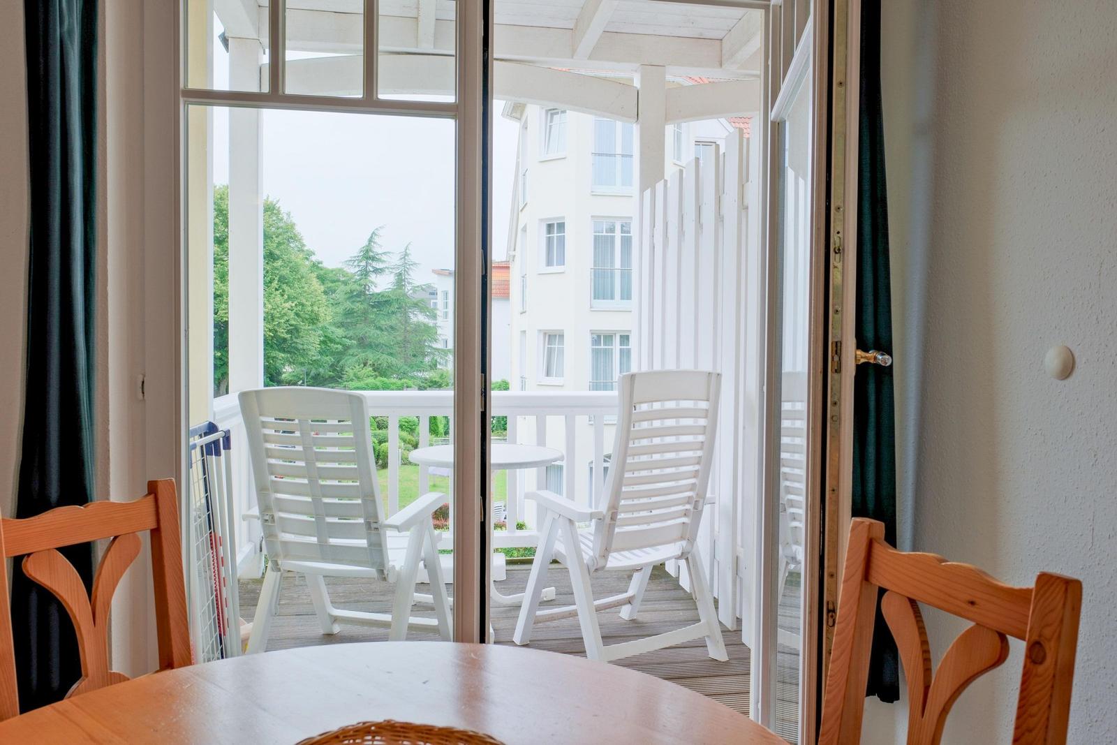 Durch Glastüren führt ein Balkon mit weißen Stühlen und Tisch. Außen sind Bäume und ein Gebäude sichtbar.