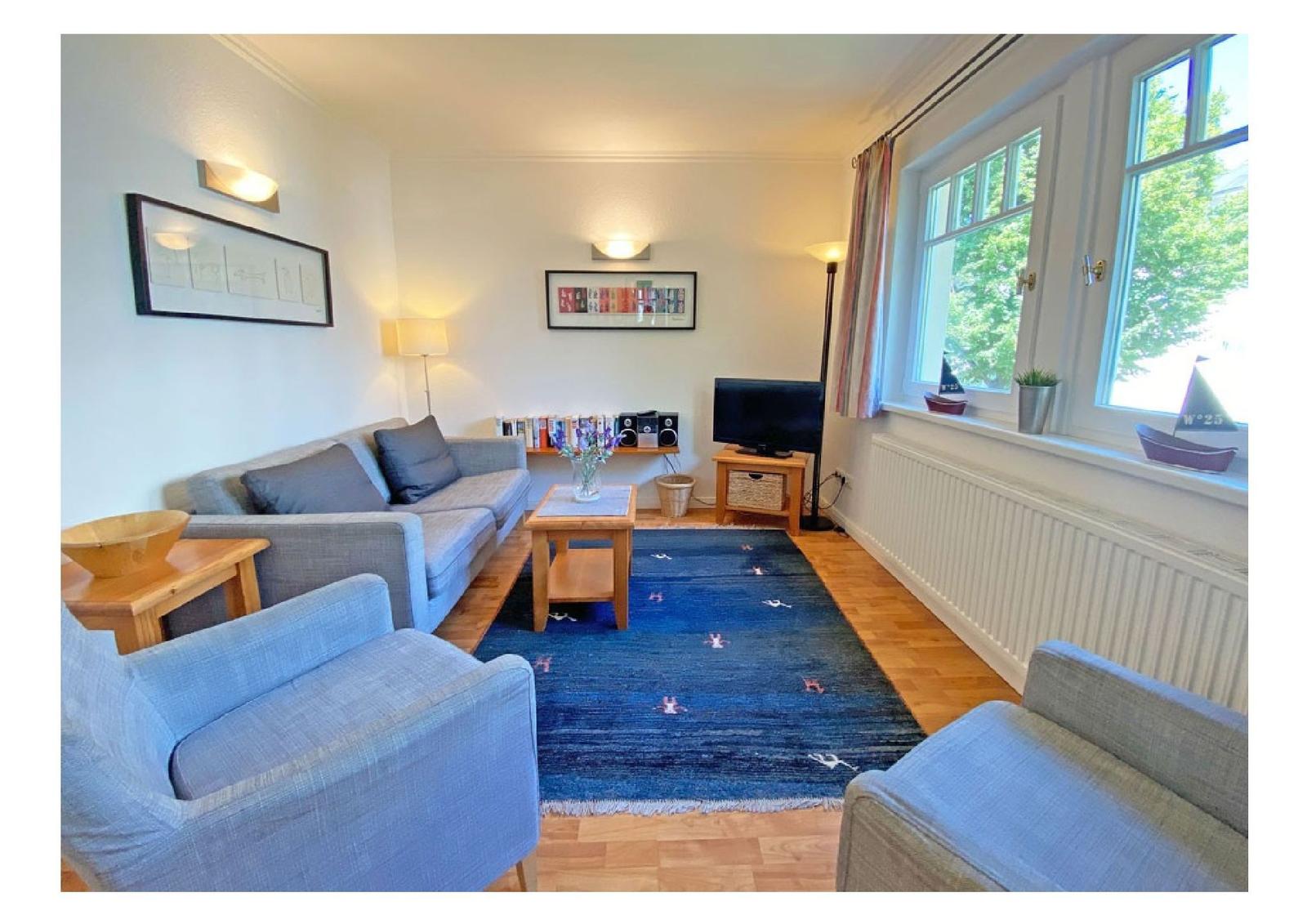 Wohnzimmer mit Sofa, Sesseln und blauem Teppich. Fenster mit Blick auf Grün.