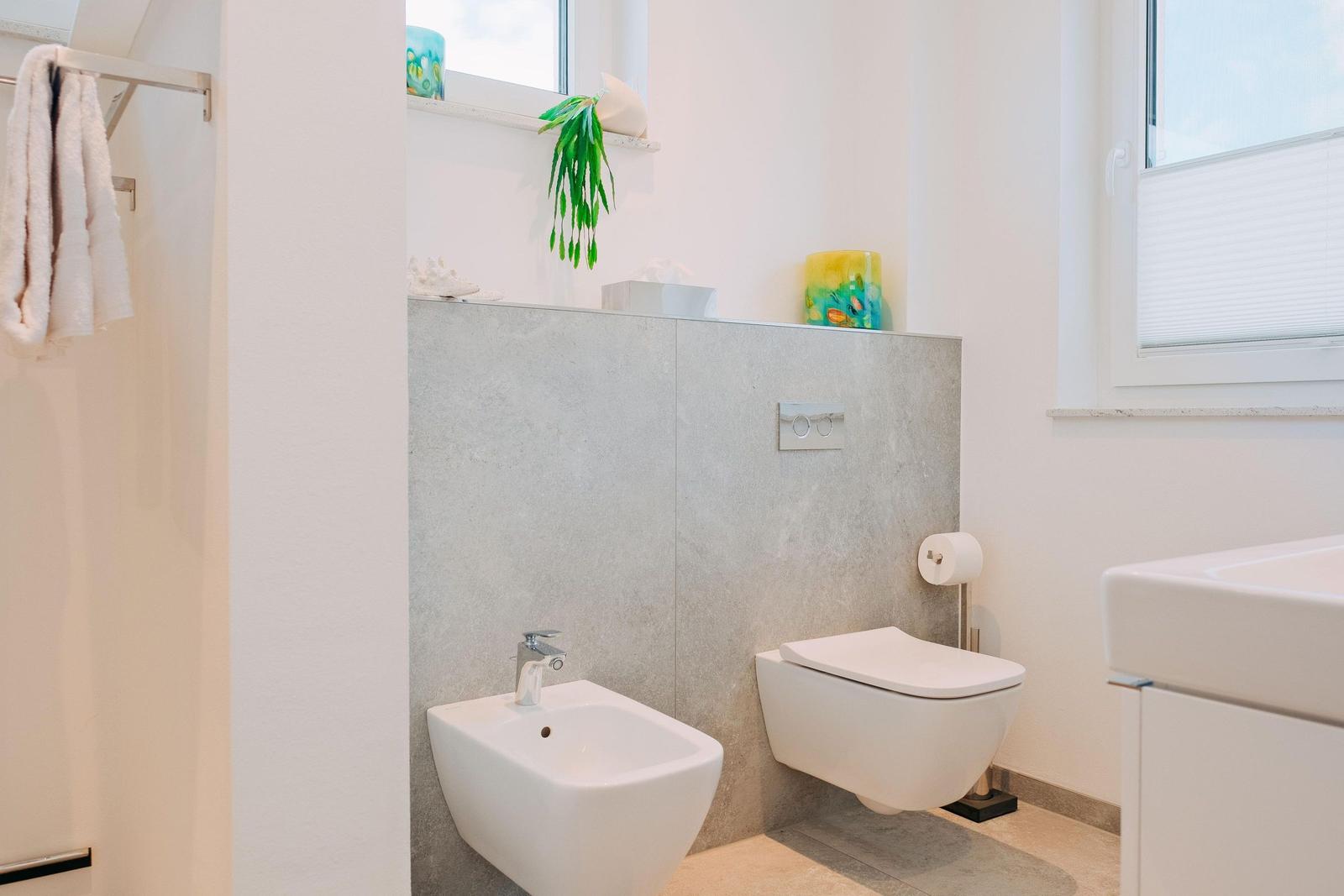 Badezimmer mit Toilette, Bidet und Waschbecken. Fenster mit Jalousie und dekorativen Gegenständen.