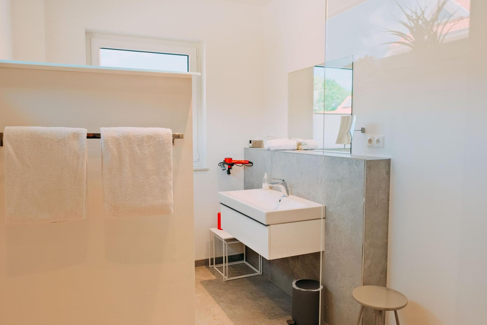 Badezimmer mit Waschbecken, Spiegel und Handtuchhalter. Fenster und kleine Sitzgelegenheit.