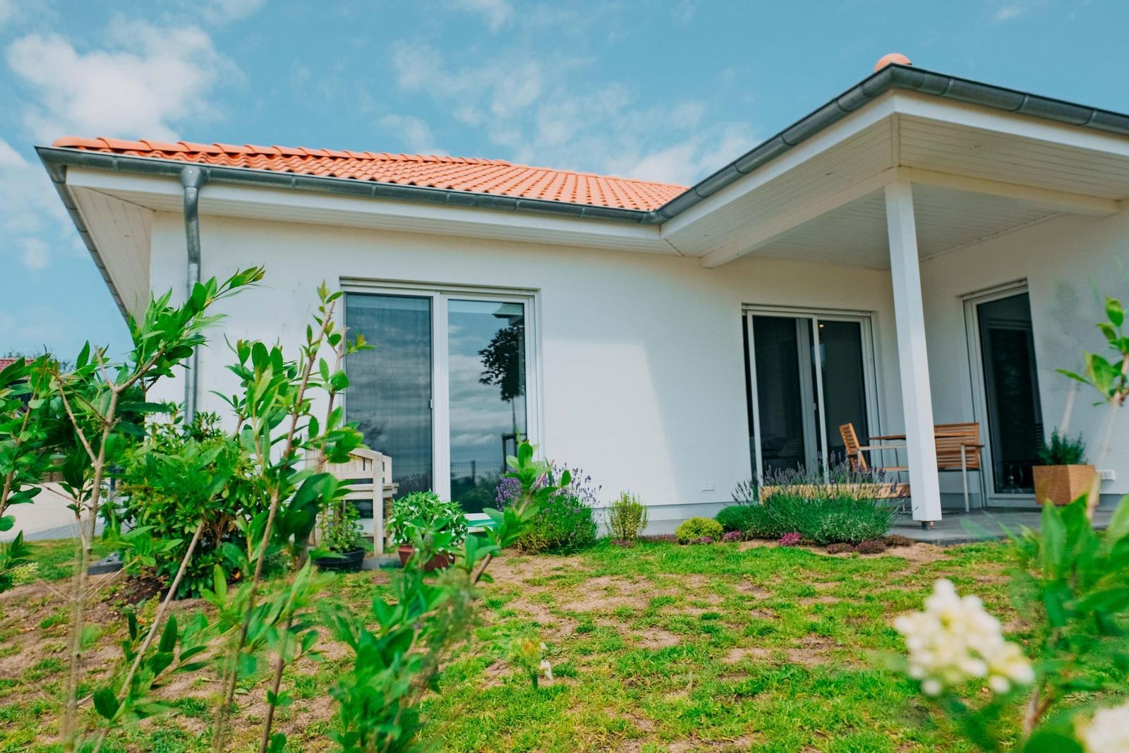 Wohnhaus mit Terrasse, Gartentüren und Garten. Weiße Fassade und rote Ziegeldach.