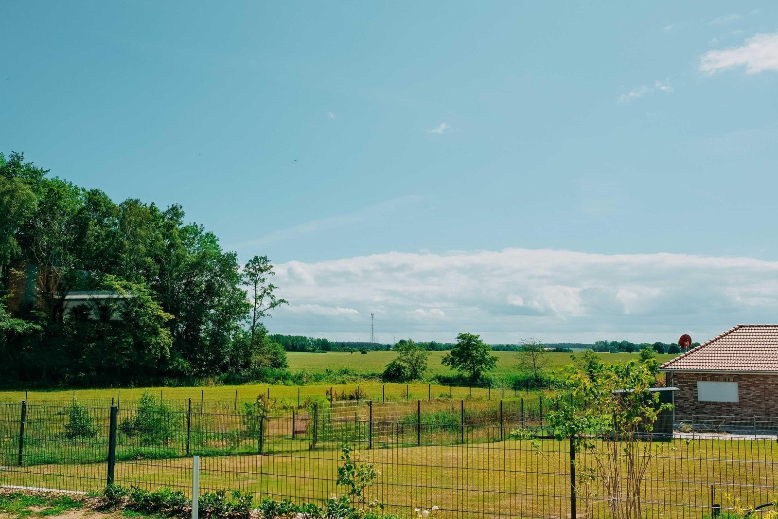 Haus mit Gartenzaun, grüne Wiesen und Bäume unter blauem Himmel.