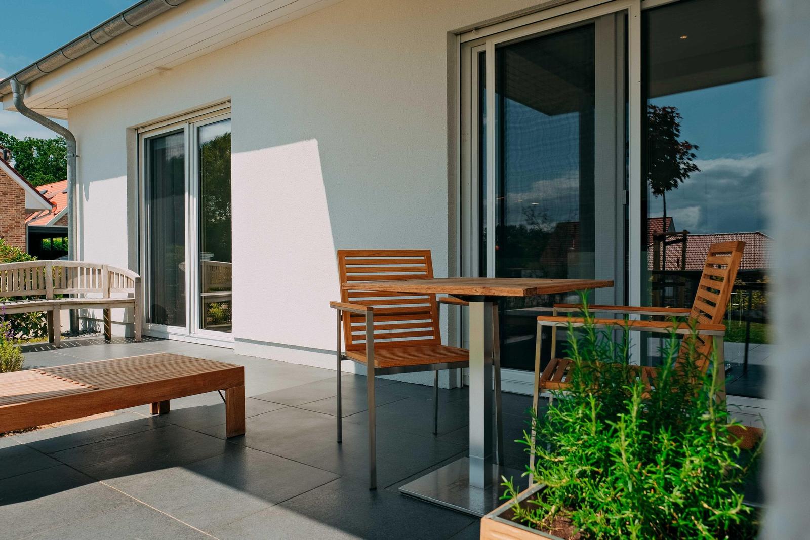 Terrasse mit Tisch, Stühlen und Liege. Glasfront und Pflanzen sind sichtbar.