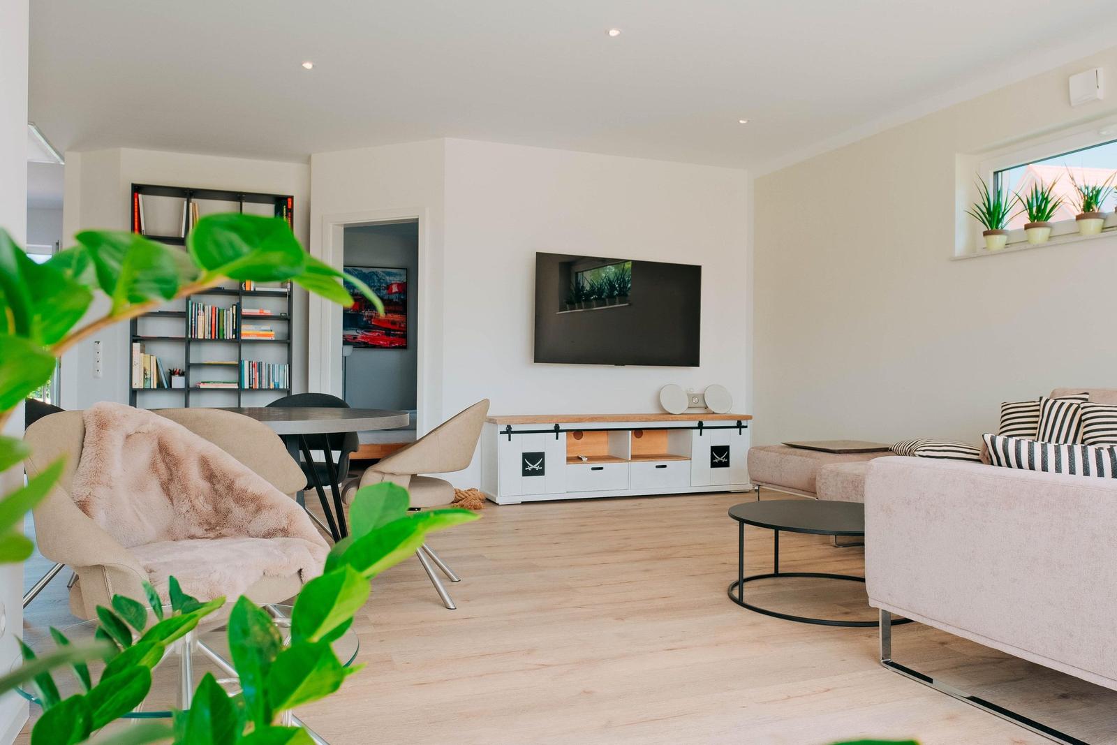 Wohnzimmer mit Sofa, Fernseher und Essbereich. Holzboden und Pflanzen sichtbar.