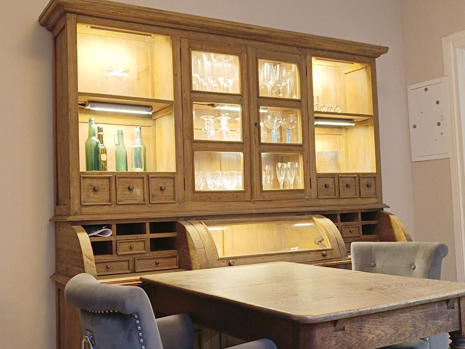 Holz-Speisekammer mit Glasfronten, Gläsern und grünen Flaschen. Holztisch und Stühle.