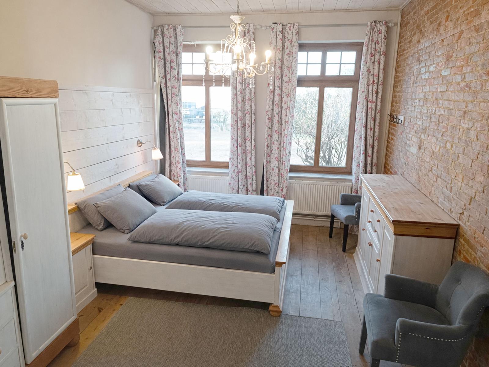Schlafzimmer mit Doppelbett, Schrank, Sessel und Fenster mit Vorhängen.