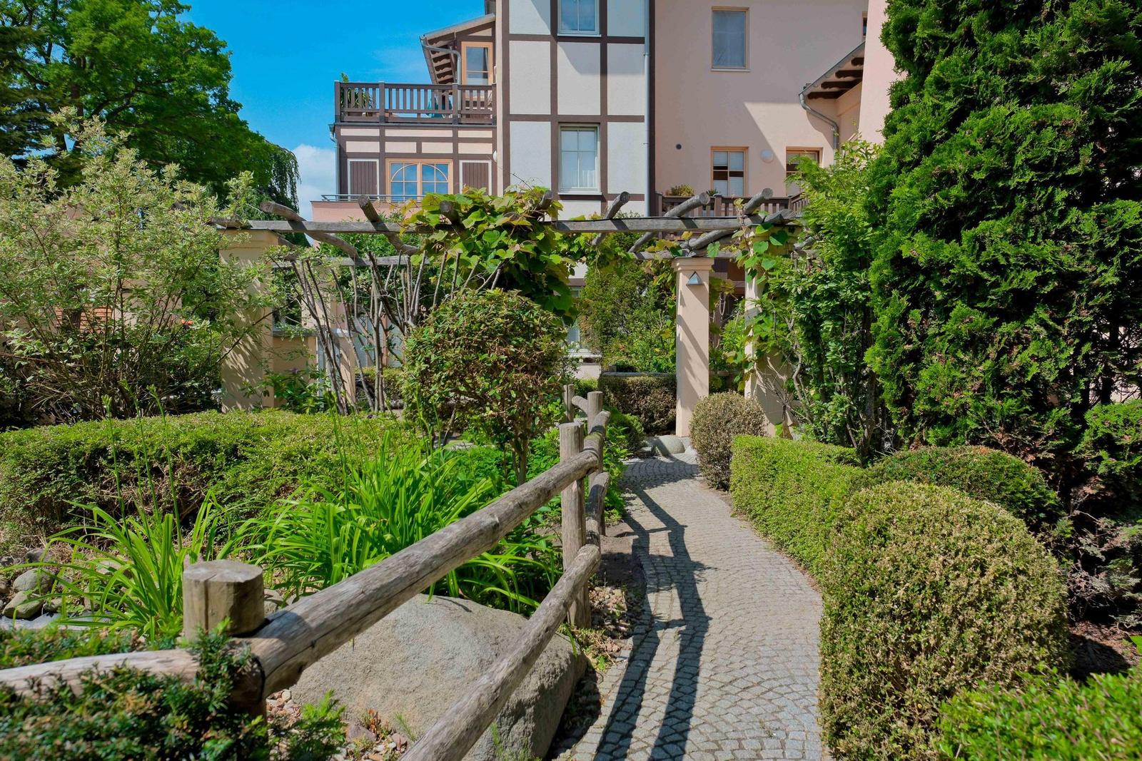 Pflasterweg durch grünen Garten mit Holzgeländer und Rankgewächsen.