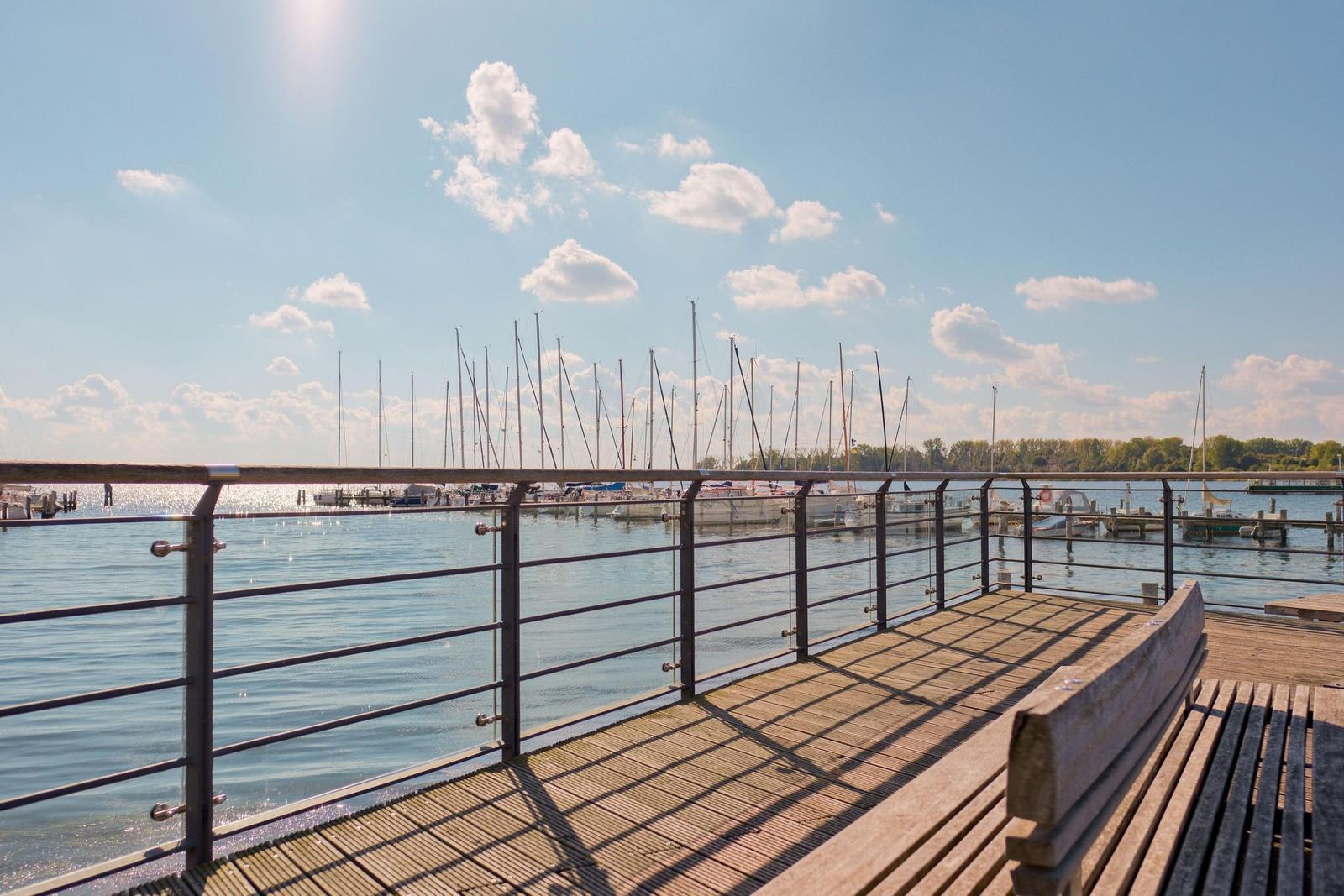 Holzterrasse mit Blick auf Hafen und Segelboote unter blauem Himmel.