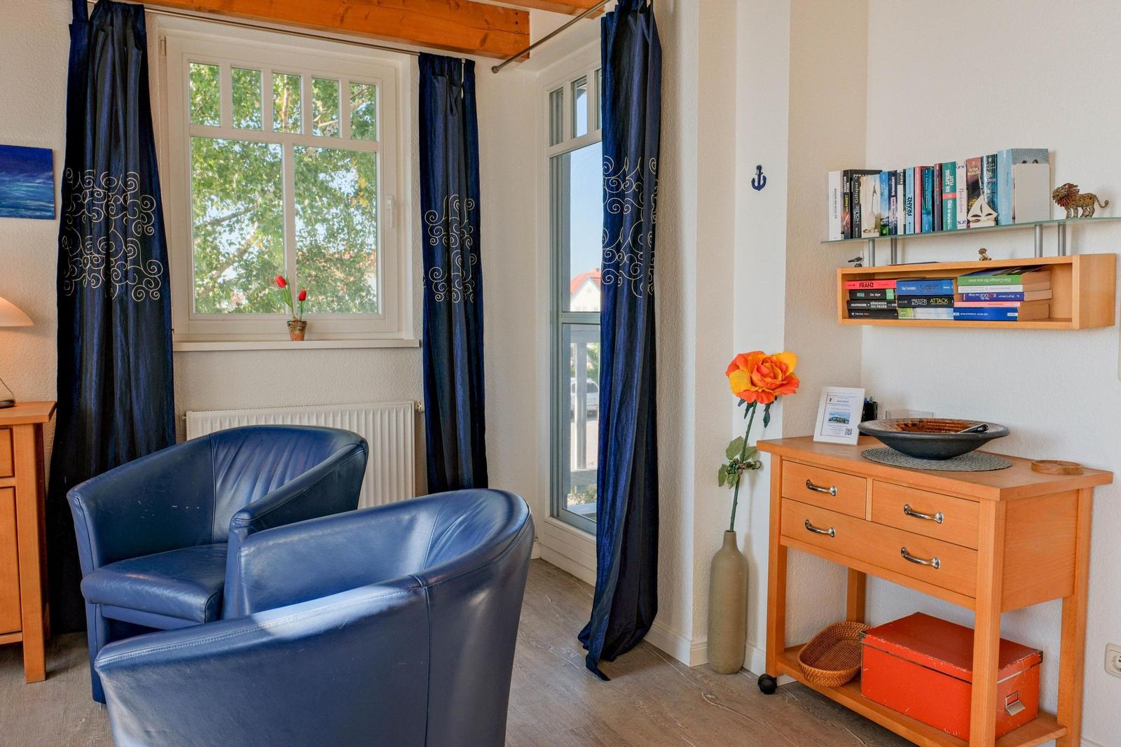 Zwei blaue Sessel stehen vor Fenstern mit dunkelblauen Vorhängen. Holzregale und ein Tisch sind sichtbar.