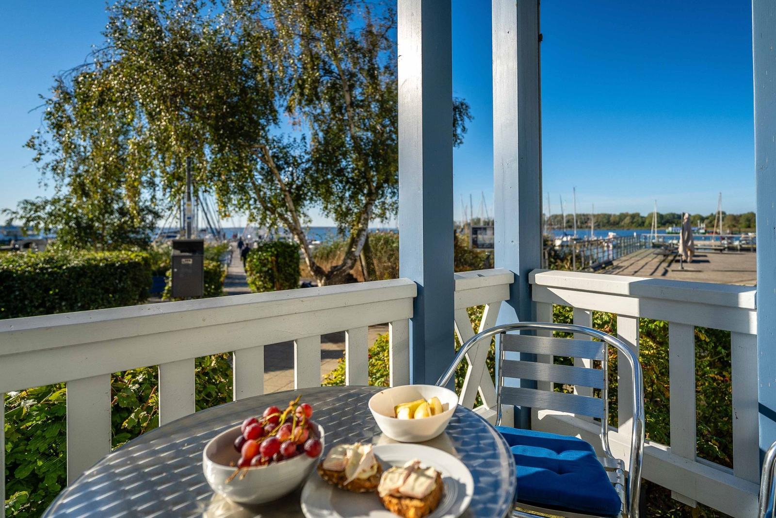 Tisch mit Obst und Brötchen auf Terrasse mit Blick auf Hafen und Bäume.
