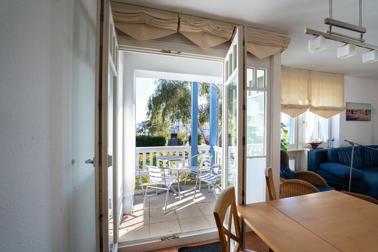 Durchgehendes Fenster mit Blick auf Terrasse mit Tisch und Stühlen. Wohnzimmer mit Sofa und Esstisch.