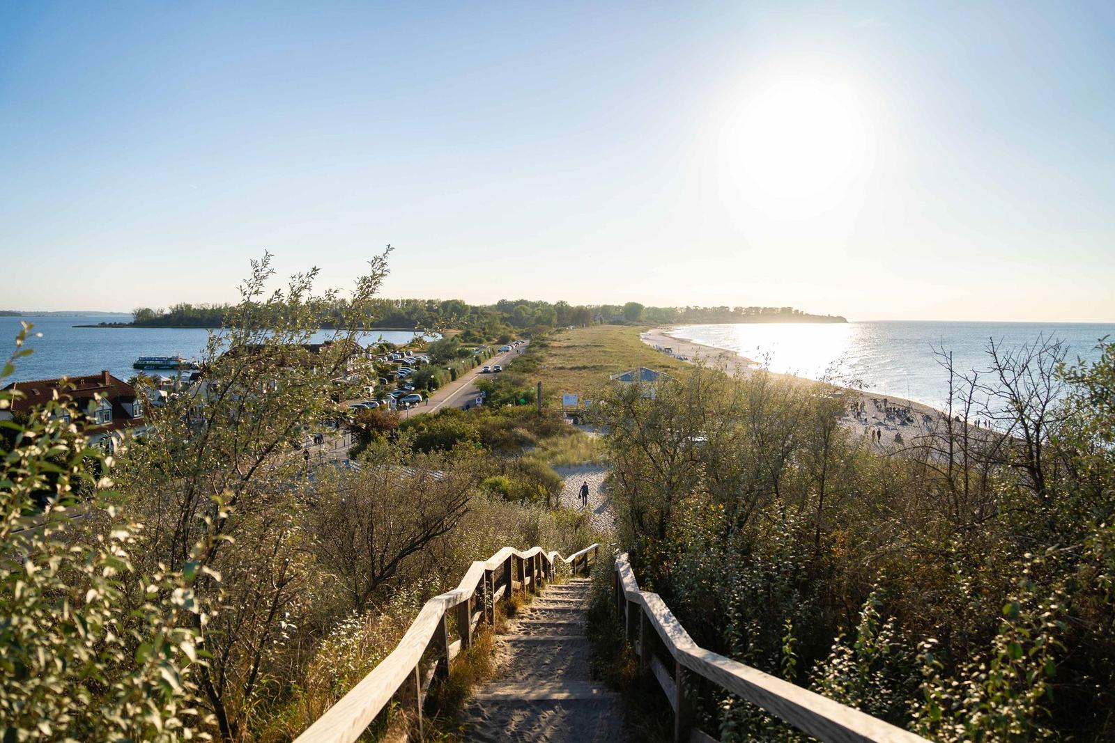 Holzsteg führt hinab zum Strand und zur Bucht. Sonne scheint, Menschen genießen die Küste.