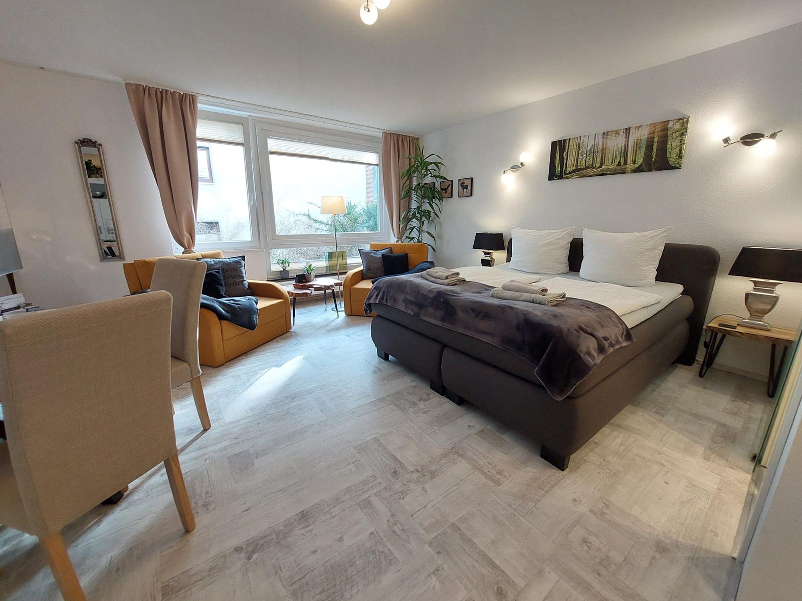 Ein Schlafzimmer mit Bett, Sessel und großem Fenster. Holzfußboden und Wandbild.