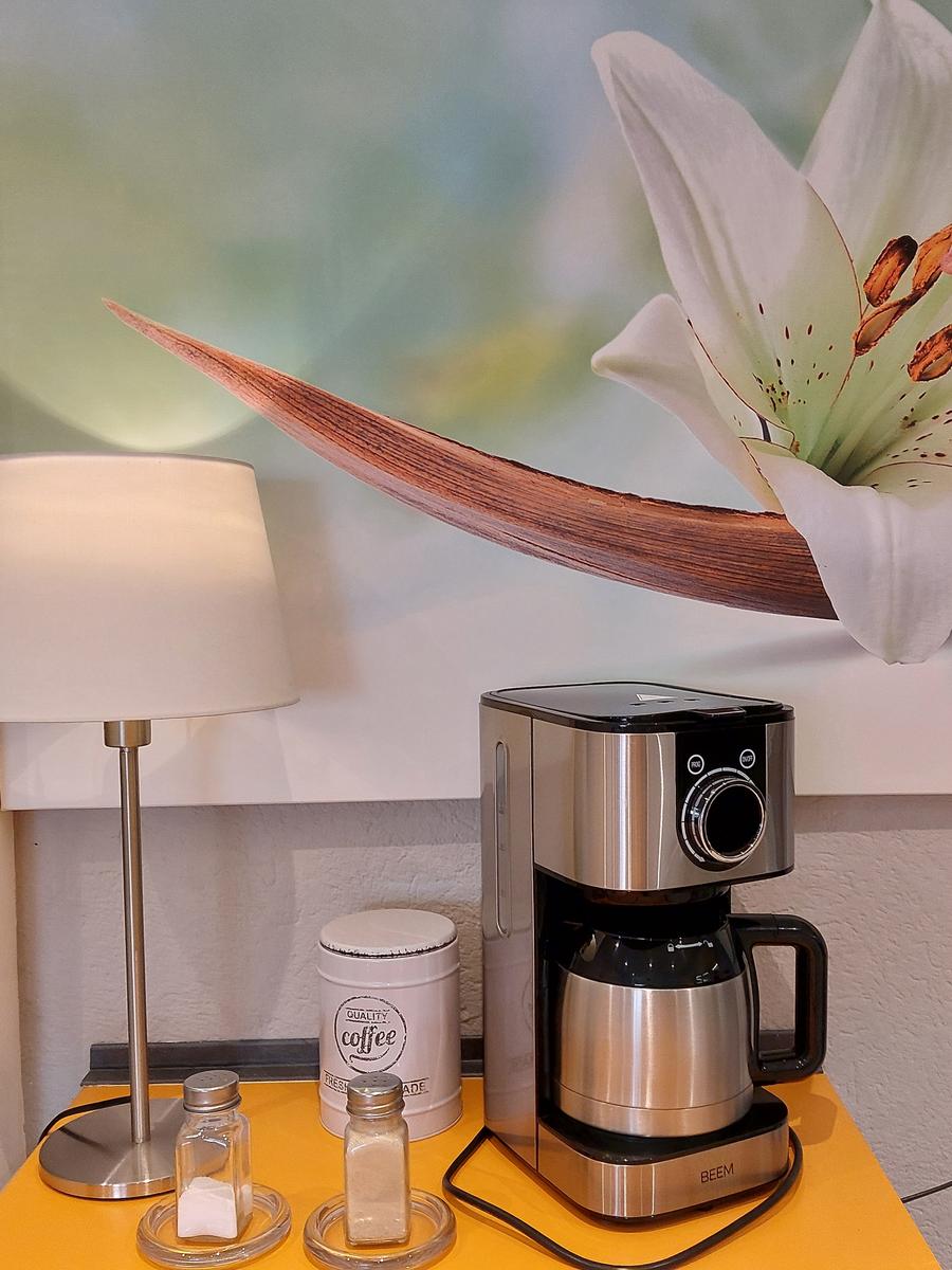 Kaffeemaschine mit Zubehör auf gelbem Tisch neben Wandbild mit Lilie.