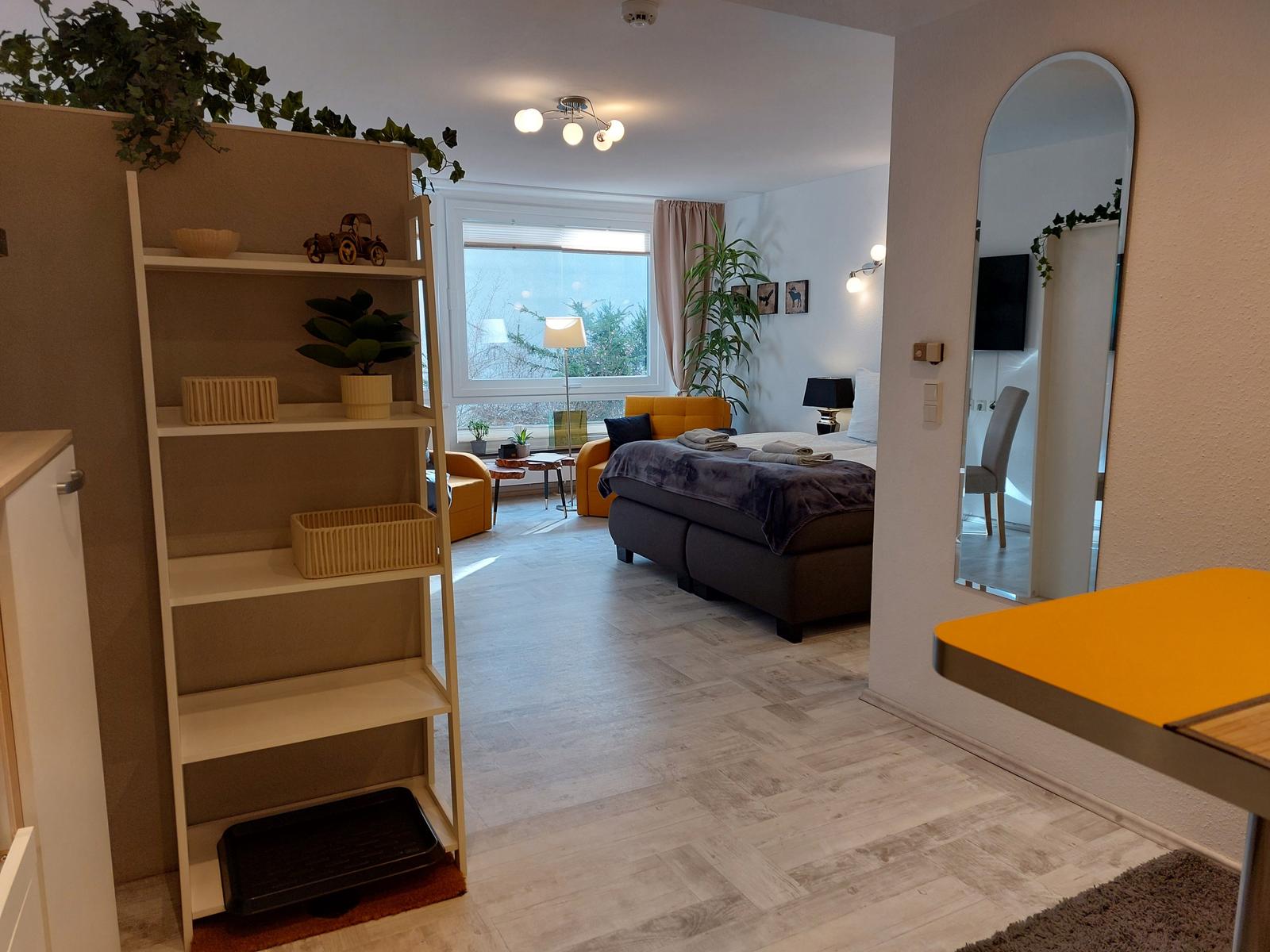 Schlafzimmer mit Bett, Sitzgelegenheit und großem Fenster. Helle Holzböden und Pflanzen.