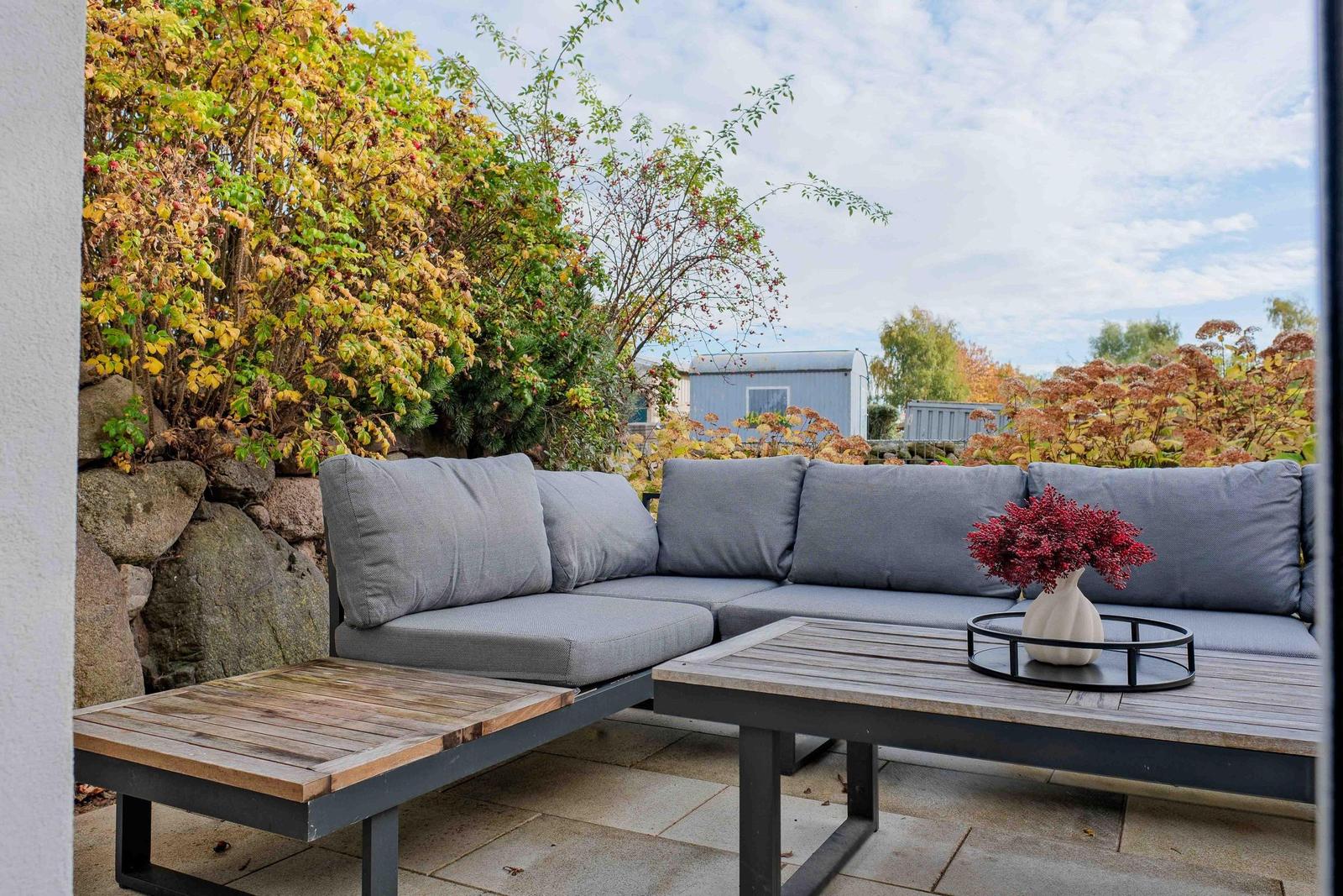 Terrasse mit grauem Sofa, Holztisch und Pflanzen im Hintergrund.
