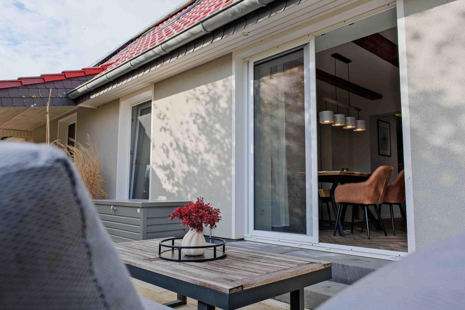 Terrasse mit Tisch und Blumen. Durch Glastür ist Esszimmer sichtbar.
