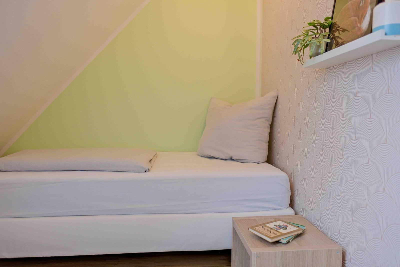 Ein Bett mit Kissen und Bettwäsche unter einer schrägen Decke. Wand mit Muster und Nachttisch.