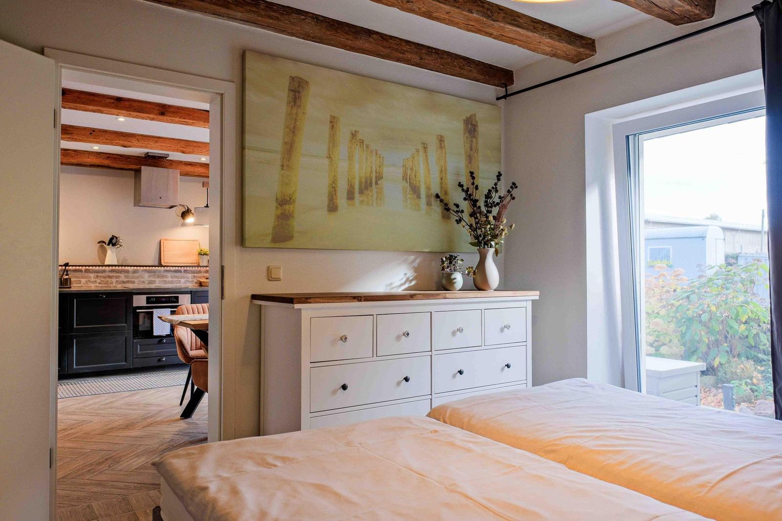 Schlafzimmer mit Blick auf Garten, weiße Kommode und Küchenbereich im Hintergrund.