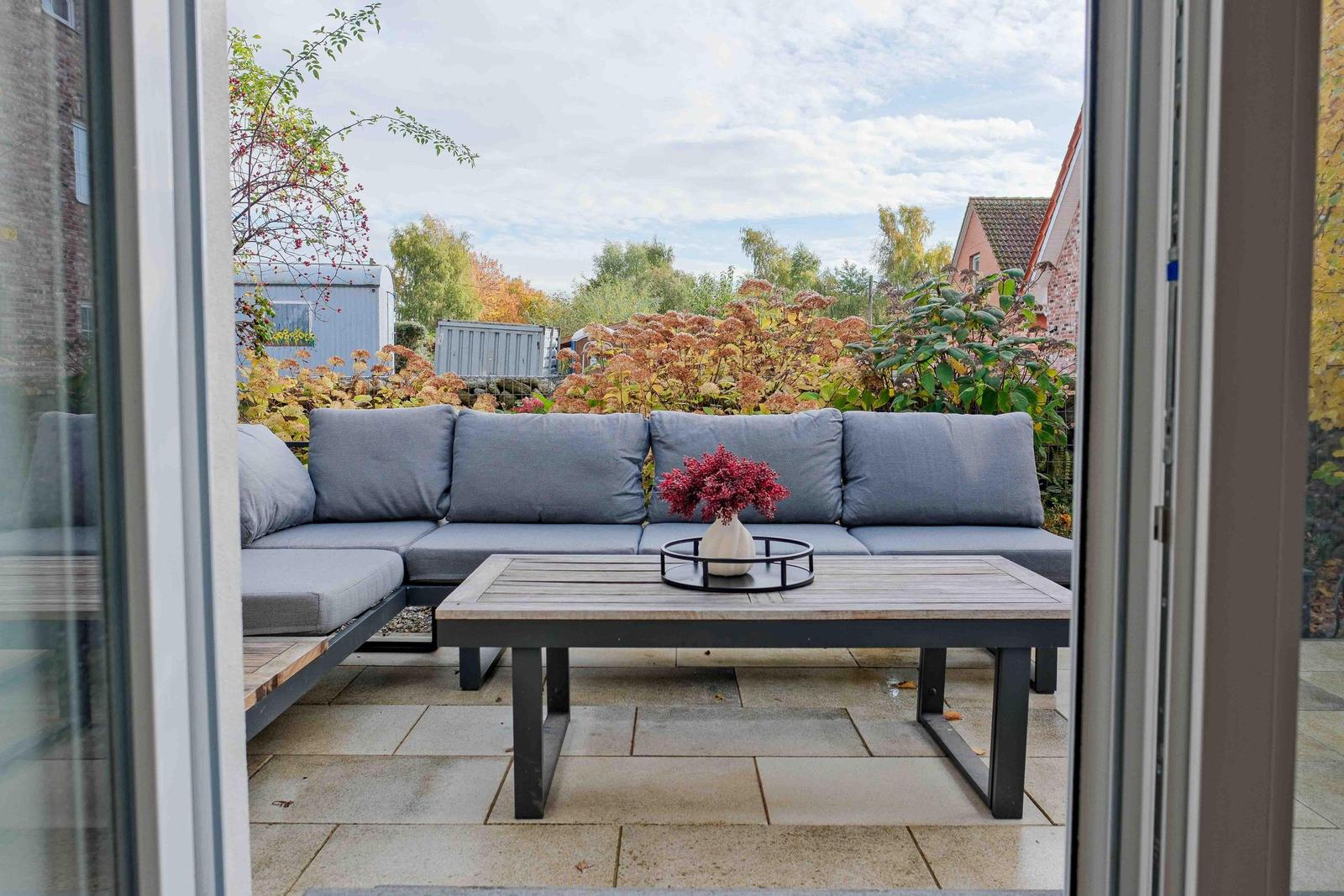 Terrasse mit grauem Sofa und Holztisch. Im Hintergrund sind Bäume und Sträucher zu sehen.