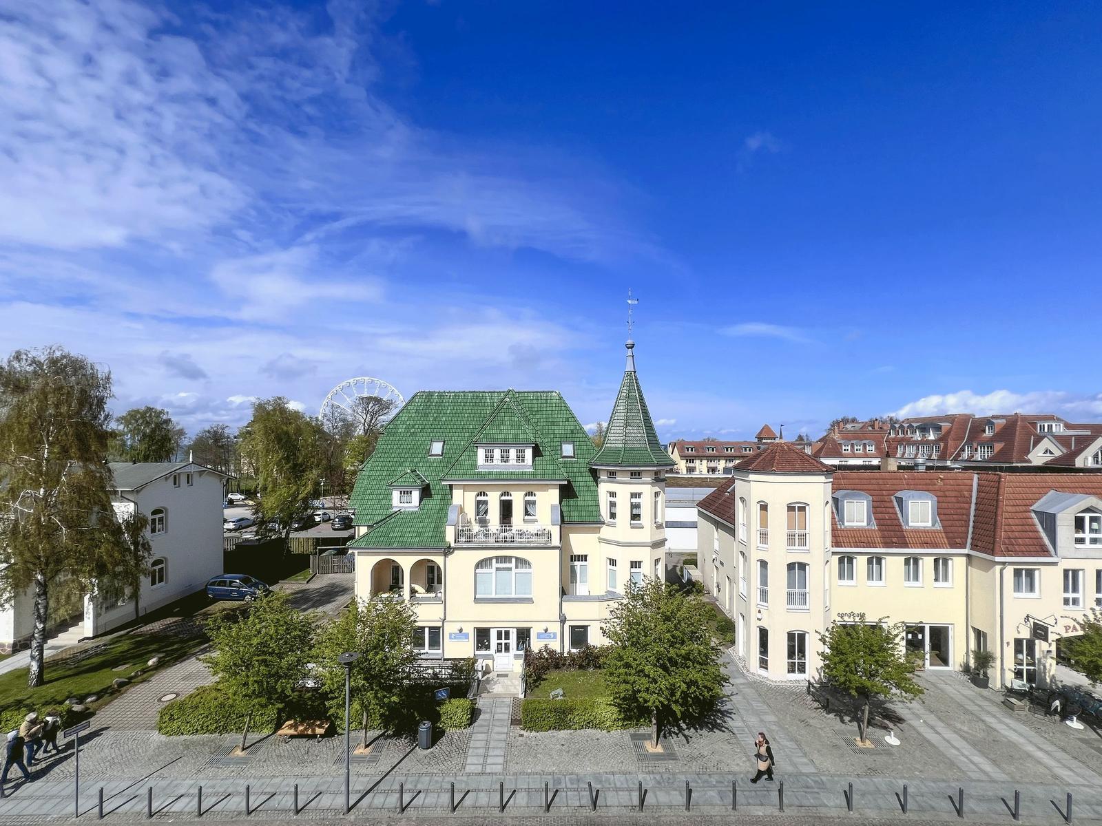 Haus mit grünem Dach und Turm in städtischer Umgebung unter blauem Himmel.