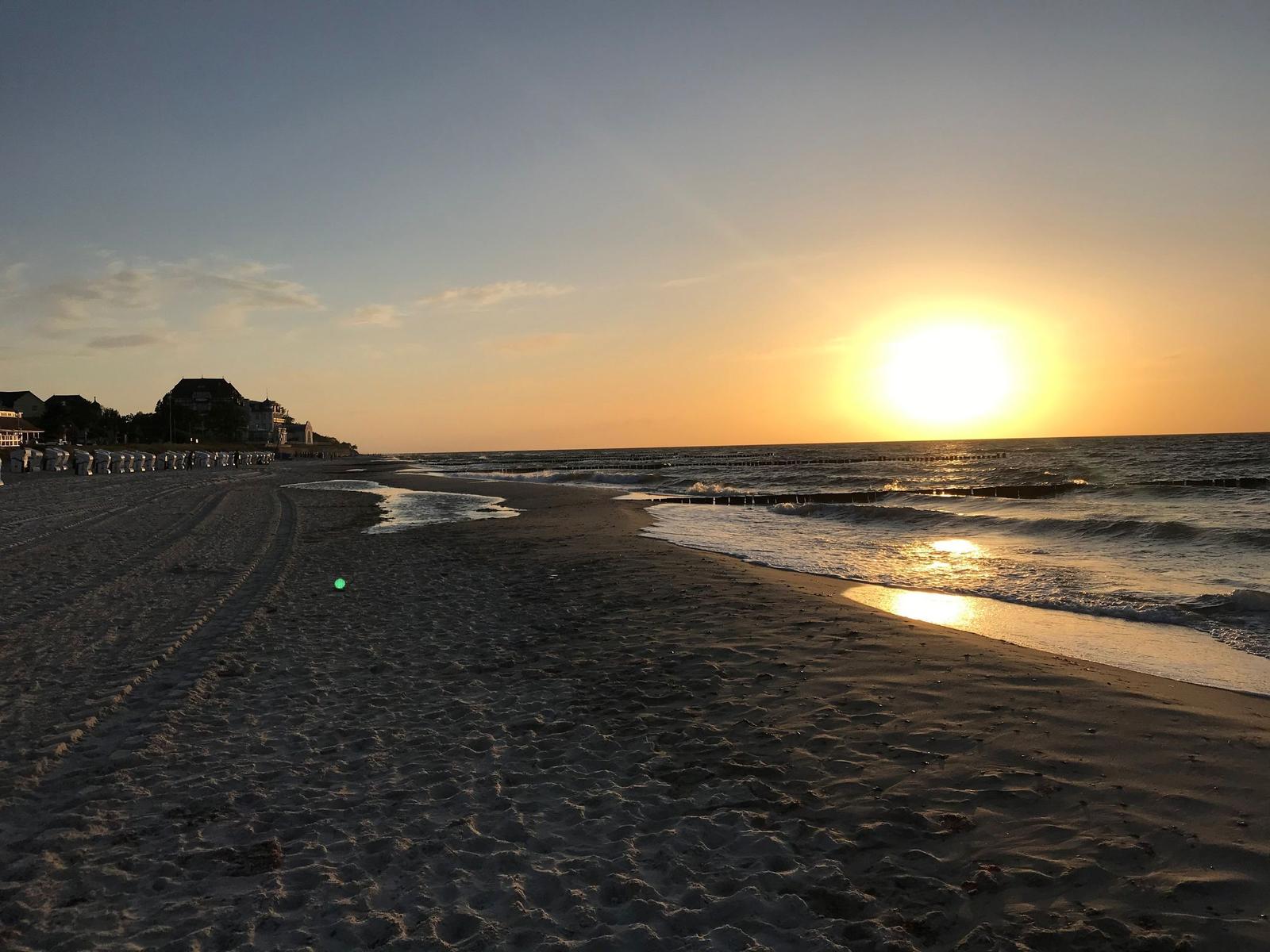 Sonnenuntergang am Strand mit Wellen und Sandstrand. Gebäude sind im Hintergrund sichtbar.
