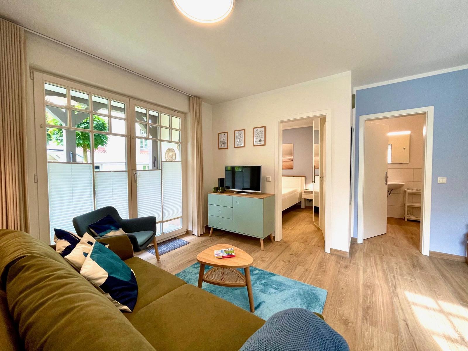 Wohnzimmer mit Sofa, Couchtisch und Blick auf Balkon. Schlafzimmer und Bad sind angebaut.
