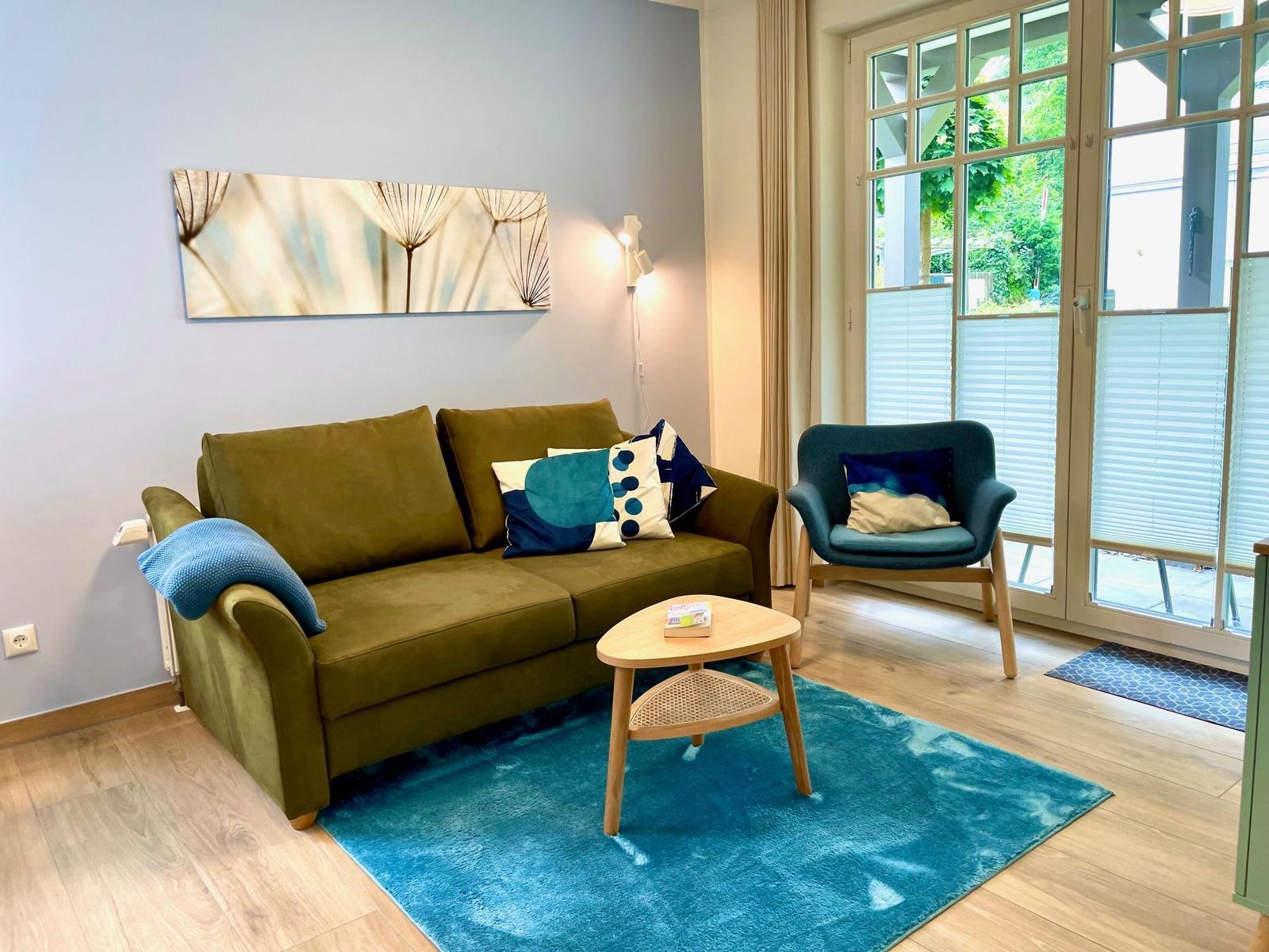 Ein Wohnzimmer mit grünem Sofa, blauem Teppich und Sitzgelegenheit vor Fenster.