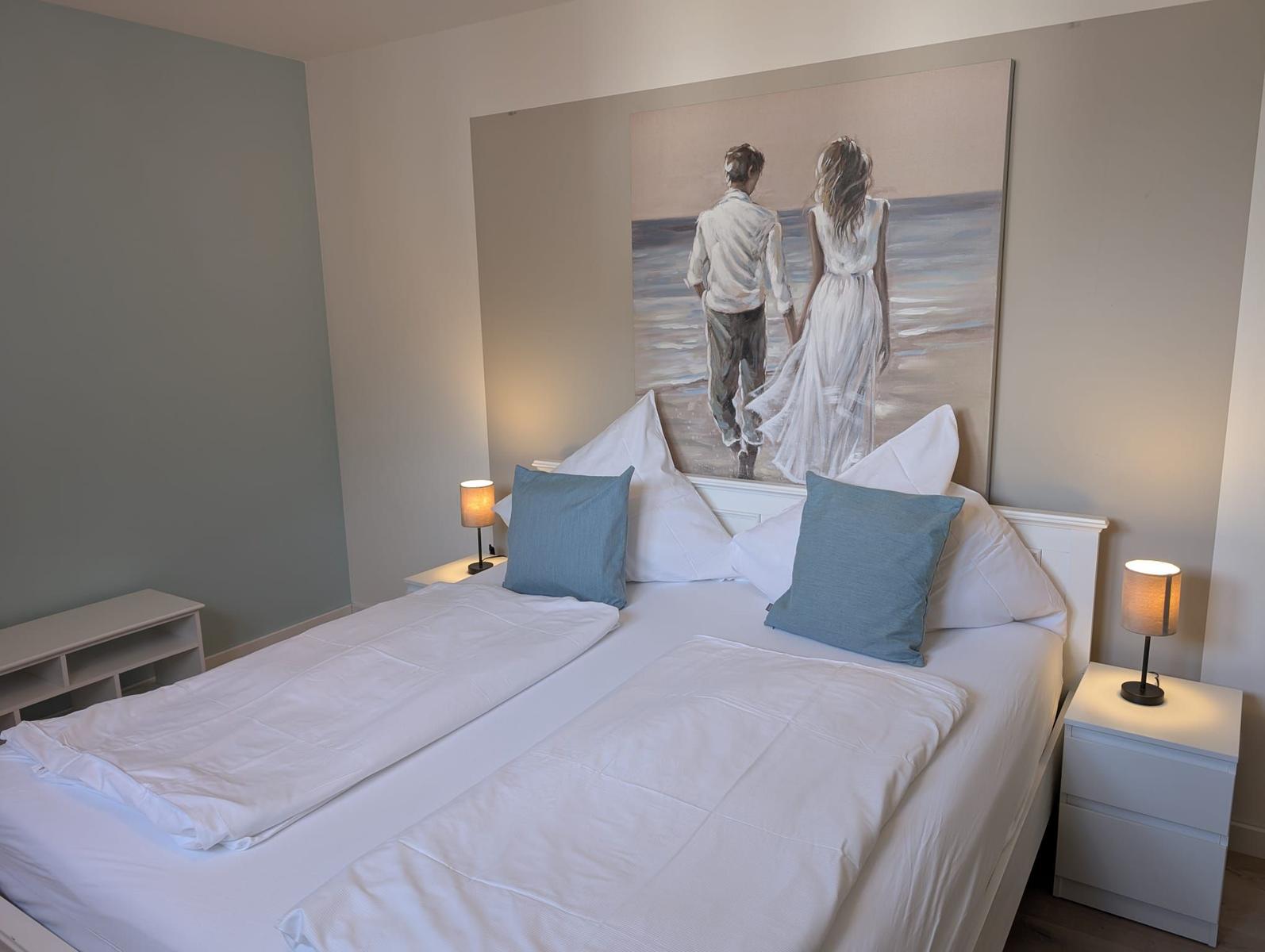Doppelbett mit weißen Bettwäsche und blauen Kissen. Wandbild über dem Bett. Zwei Nachttische mit Lampen.