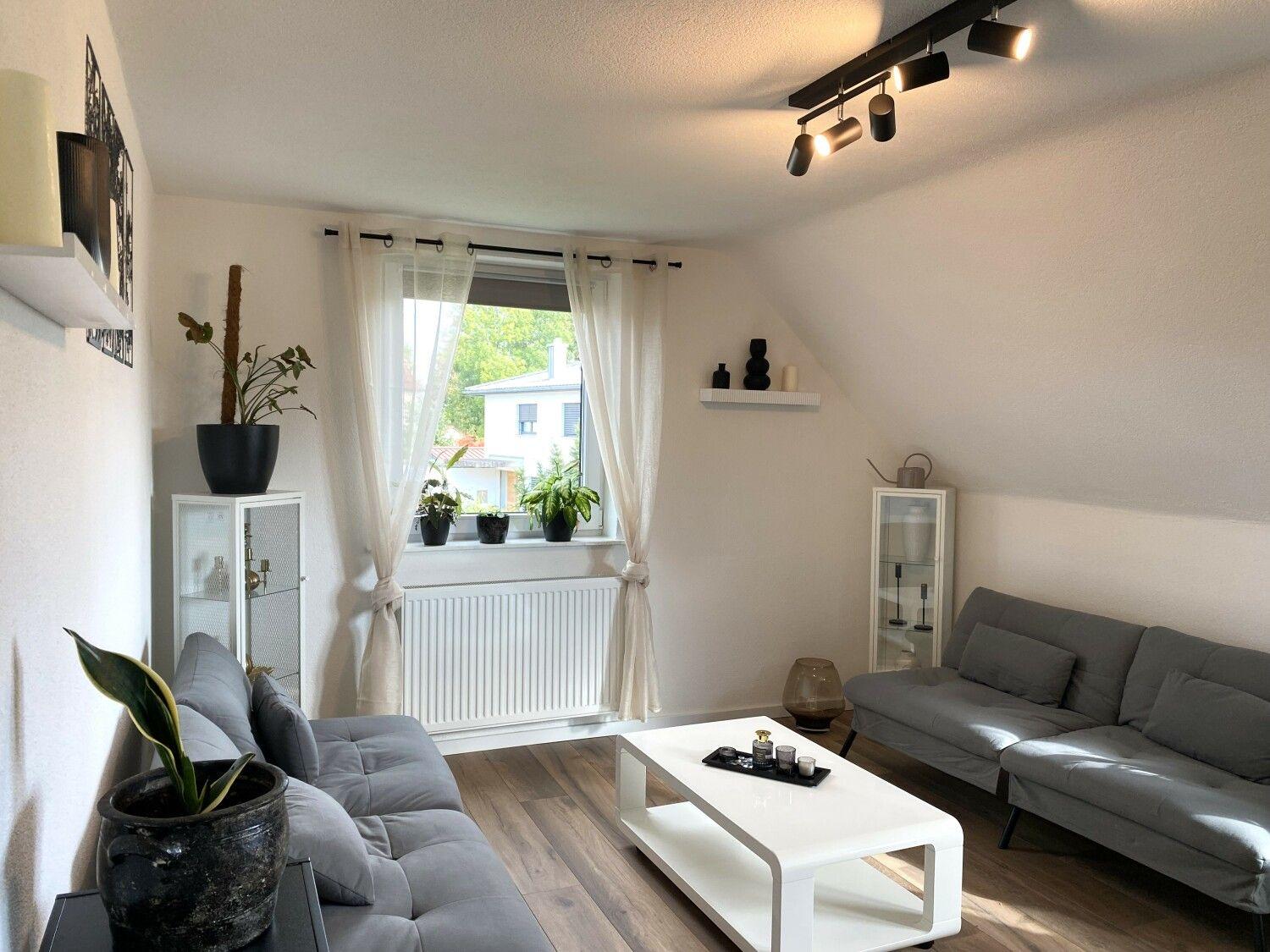 Wohnzimmer mit grauem Sofa, weißem Tisch und Pflanzen an Fenster.