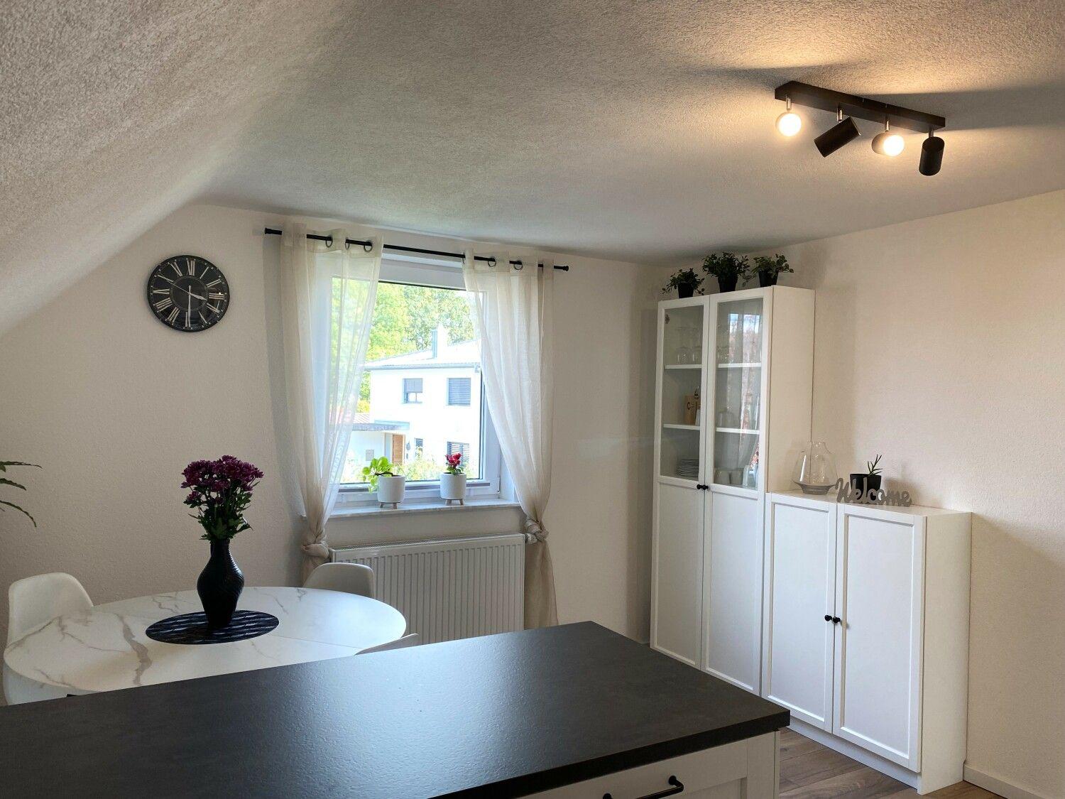 Esszimmer mit Tisch, Schrank und Fenster. Decke geneigt, Wanduhr und Pflanzen.