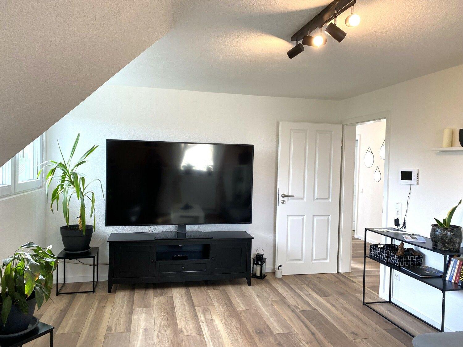 Wohnzimmer mit Fernseher, Pflanzen und Holzboden. Tür führt in andere Räume.