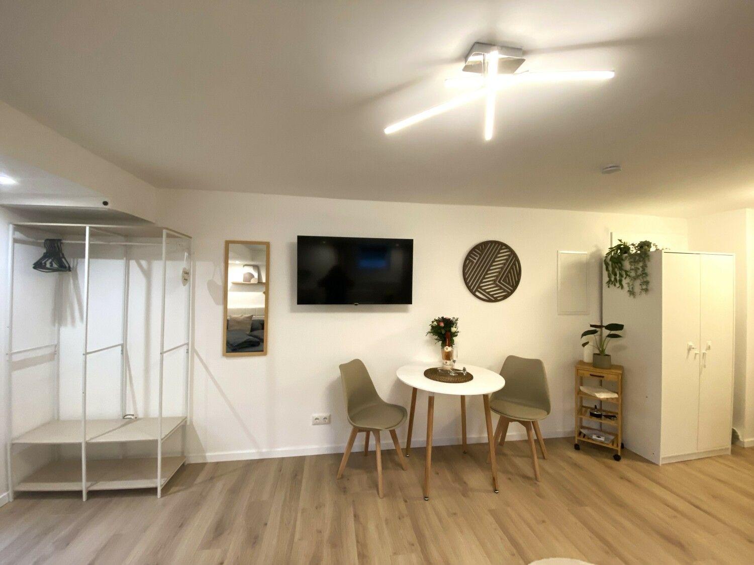 Wohnraum mit Esstisch, TV und Kleiderschrank. Holzfußboden und weiße Wände.