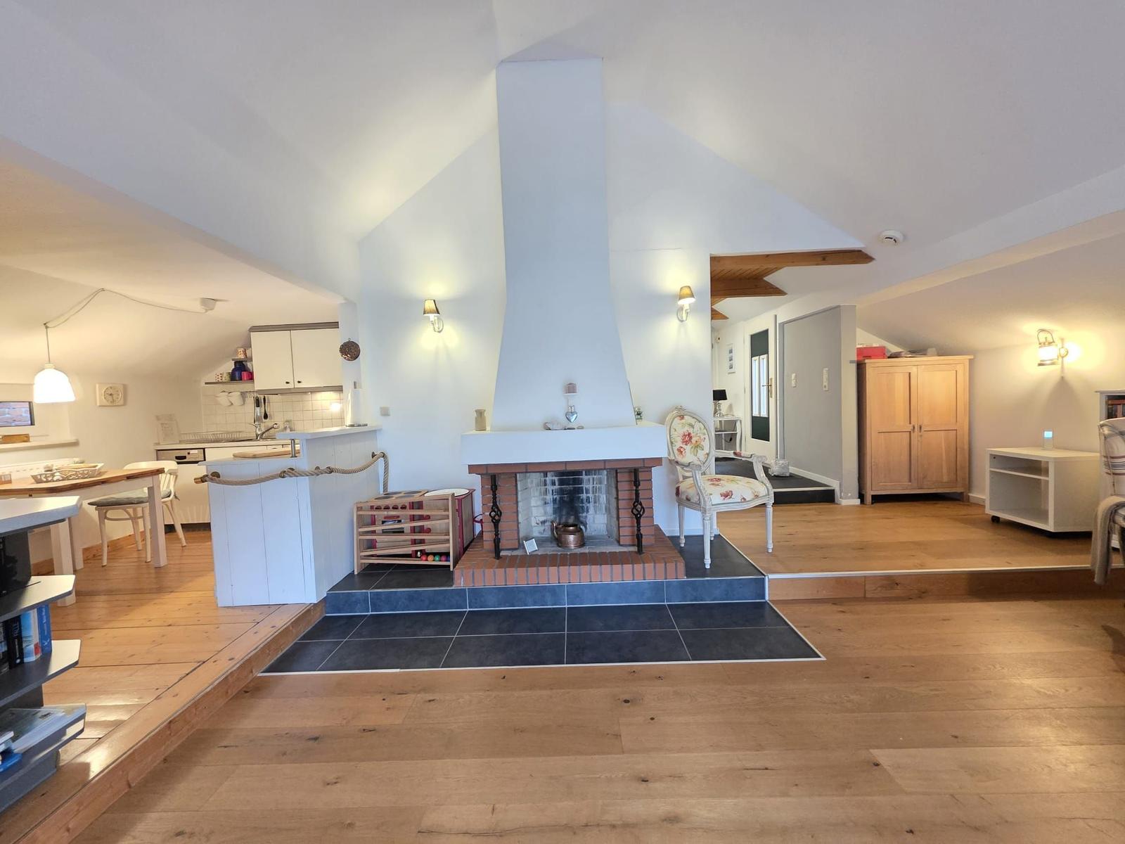Living area with fireplace, wooden floor, and kitchen area in background.
