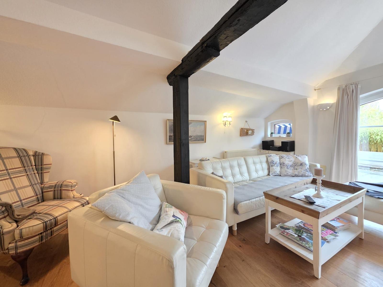 Living room with sofa, armchair, and wooden table. Visible beams and window.
