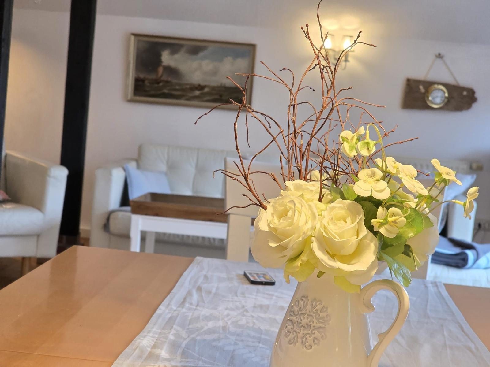 Table with flower vase, behind it living area with sofa and wall picture.