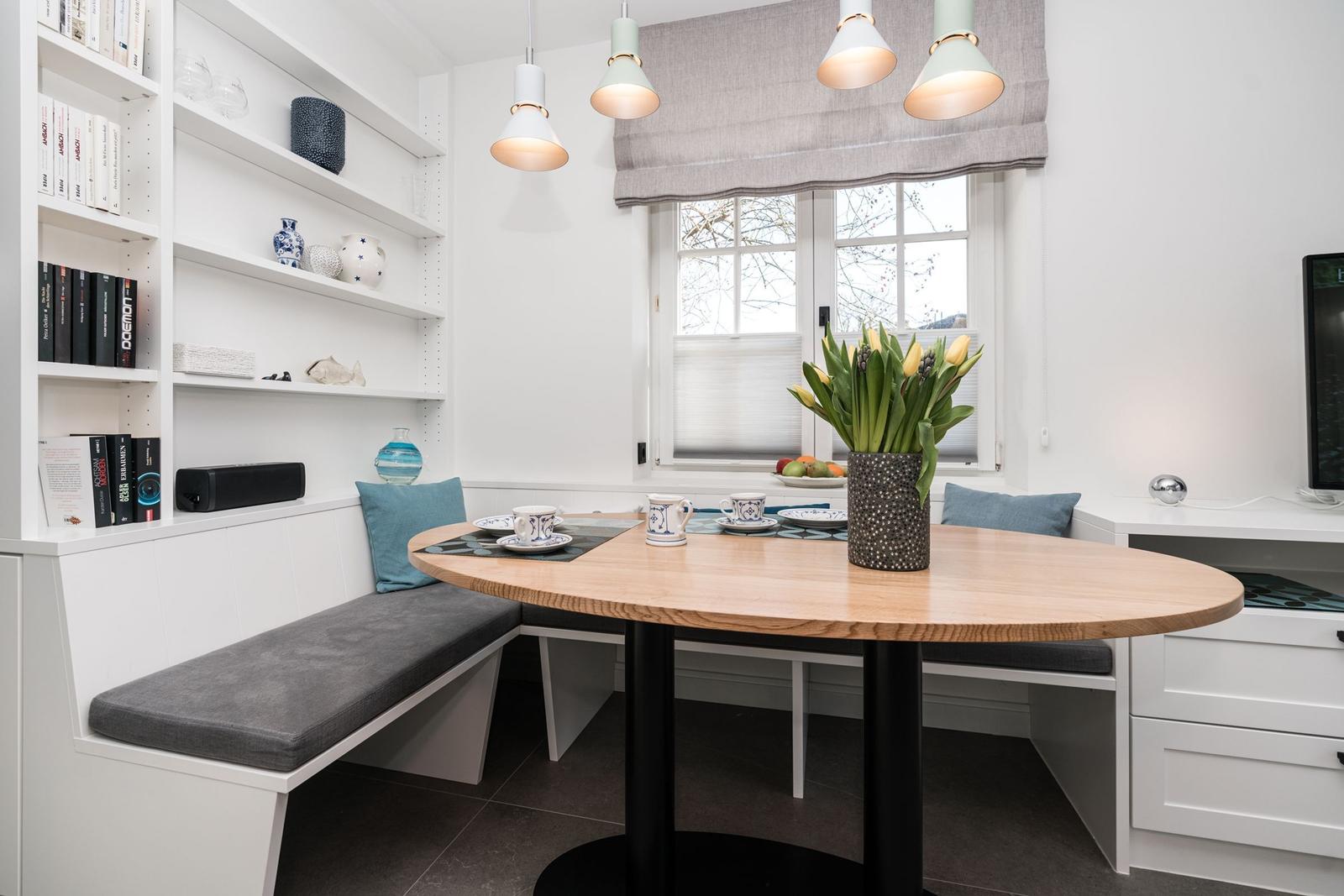 Rundtisch mit Sitzbank, Bücherregal und Fenster mit Blumen.