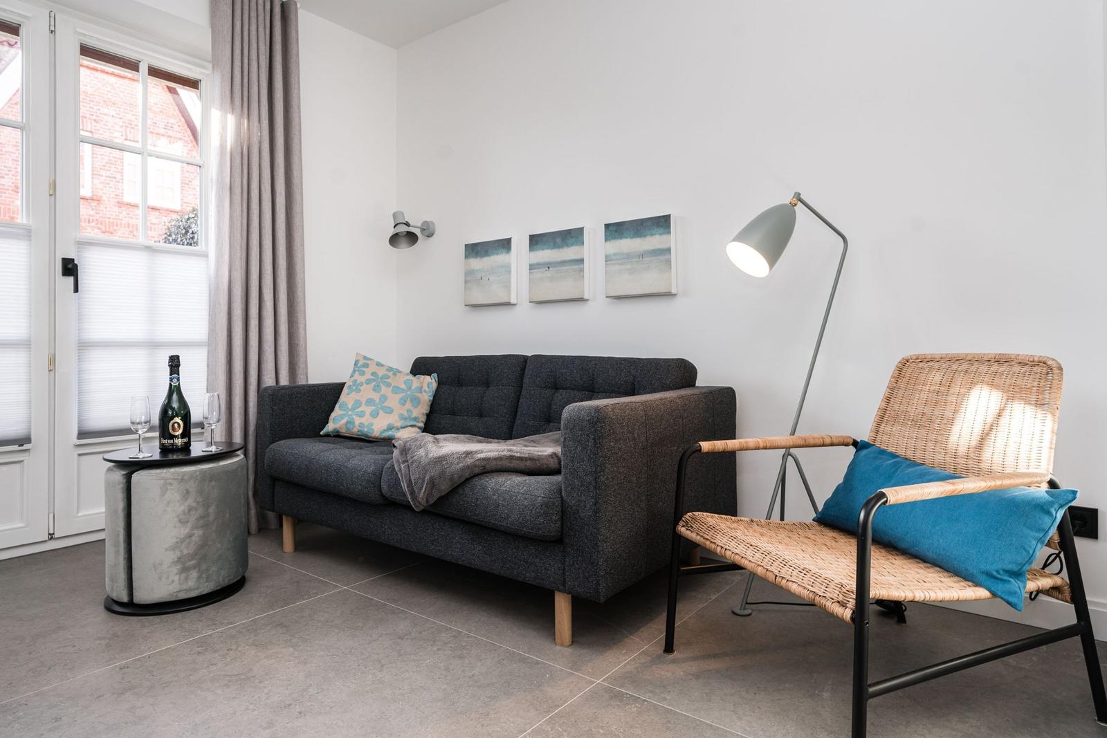 Ein Wohnzimmer mit grauem Sofa, Stuhl und Fenster. Auf Tisch steht eine Flasche.