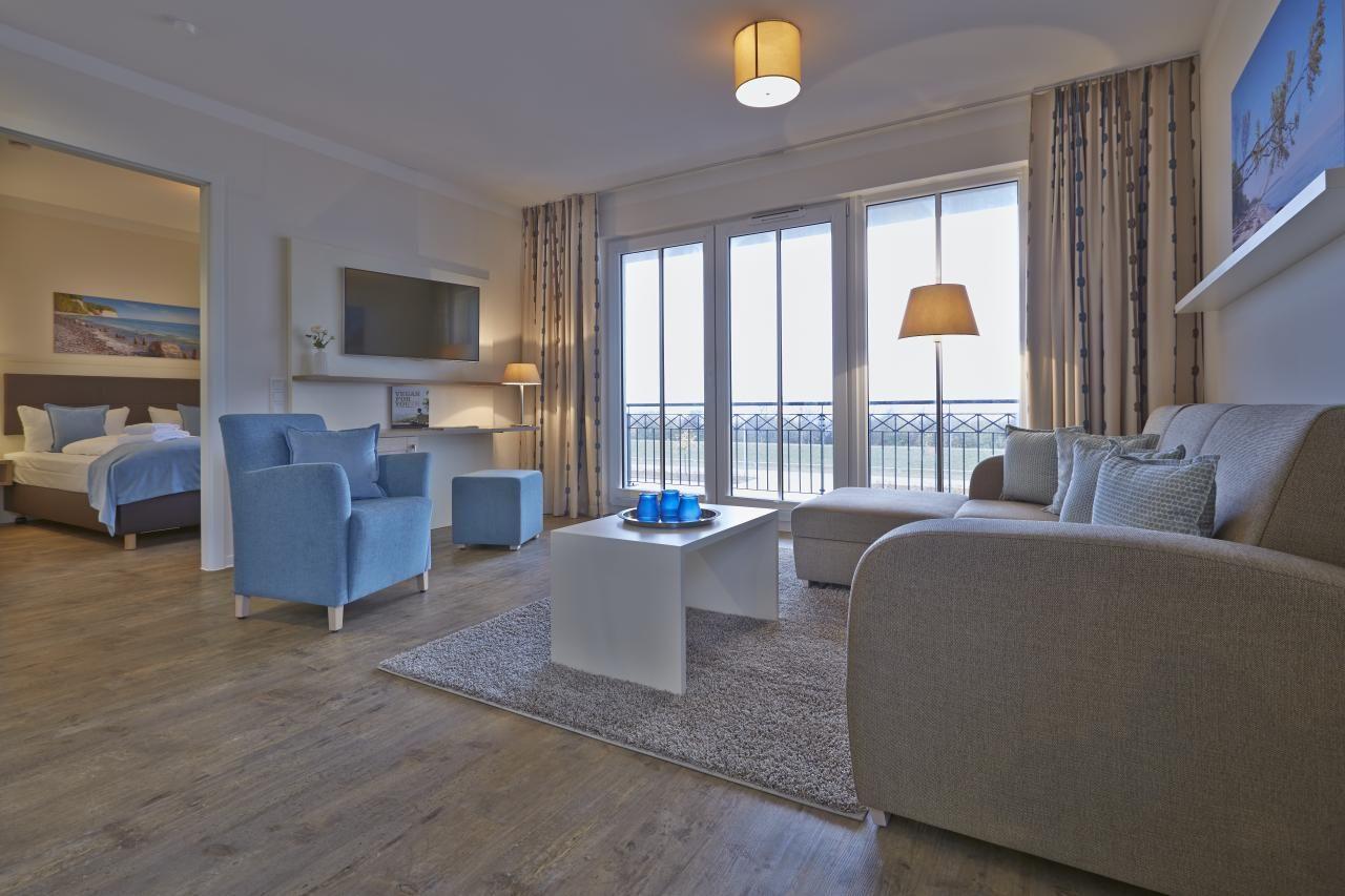 Wohnbereich mit Sofa, Sessel und Balkon. Schlafzimmer im Hintergrund.