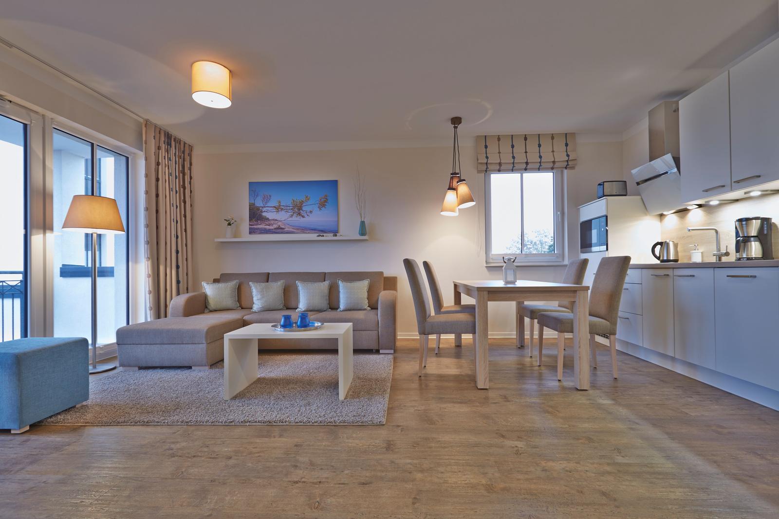 Wohnbereich mit Sofa, Esstisch und Küchenzeile. Holzboden und große Fenster.