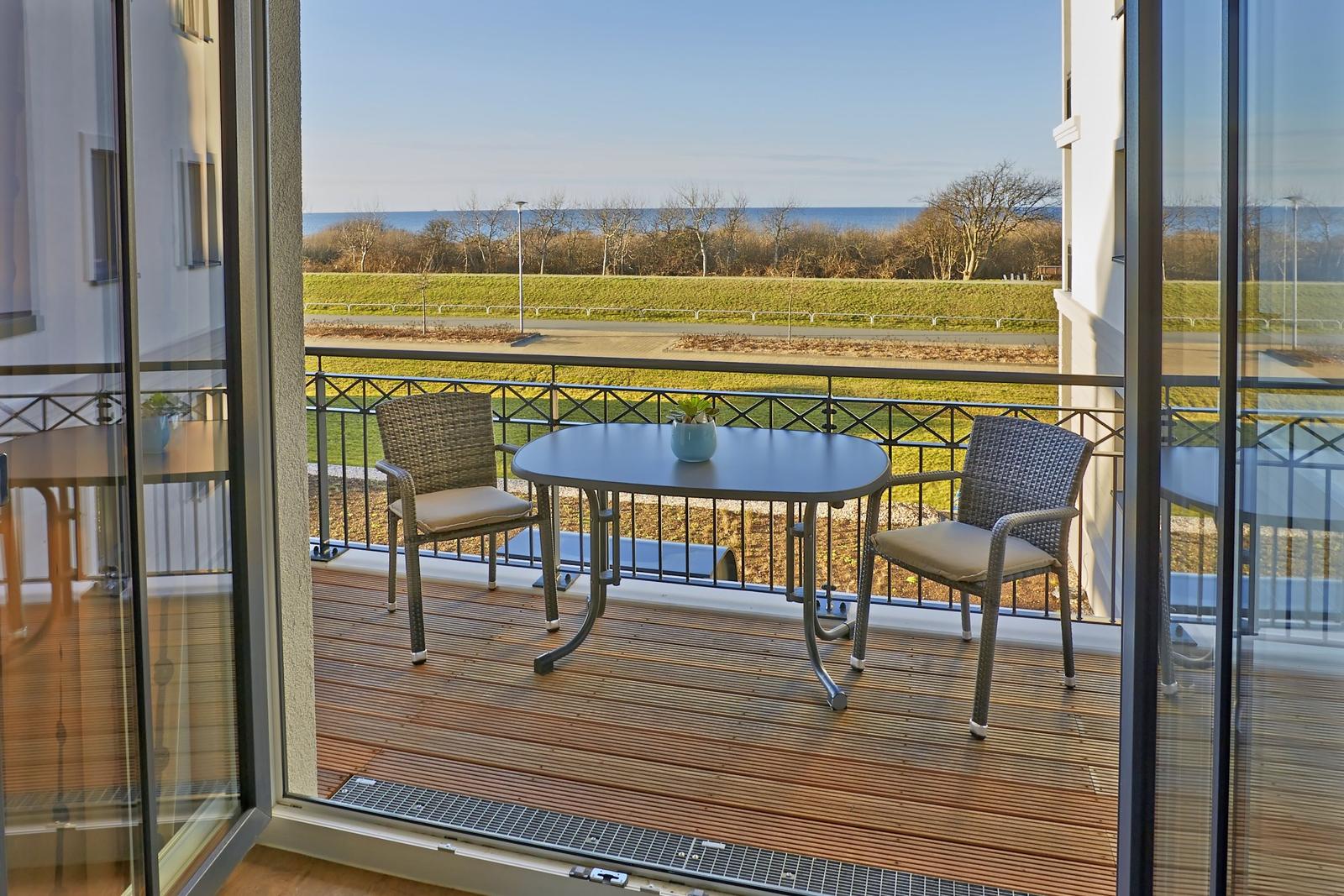 Terrasse mit Tisch und zwei Stühlen, Blick auf Grünfläche und Meer.